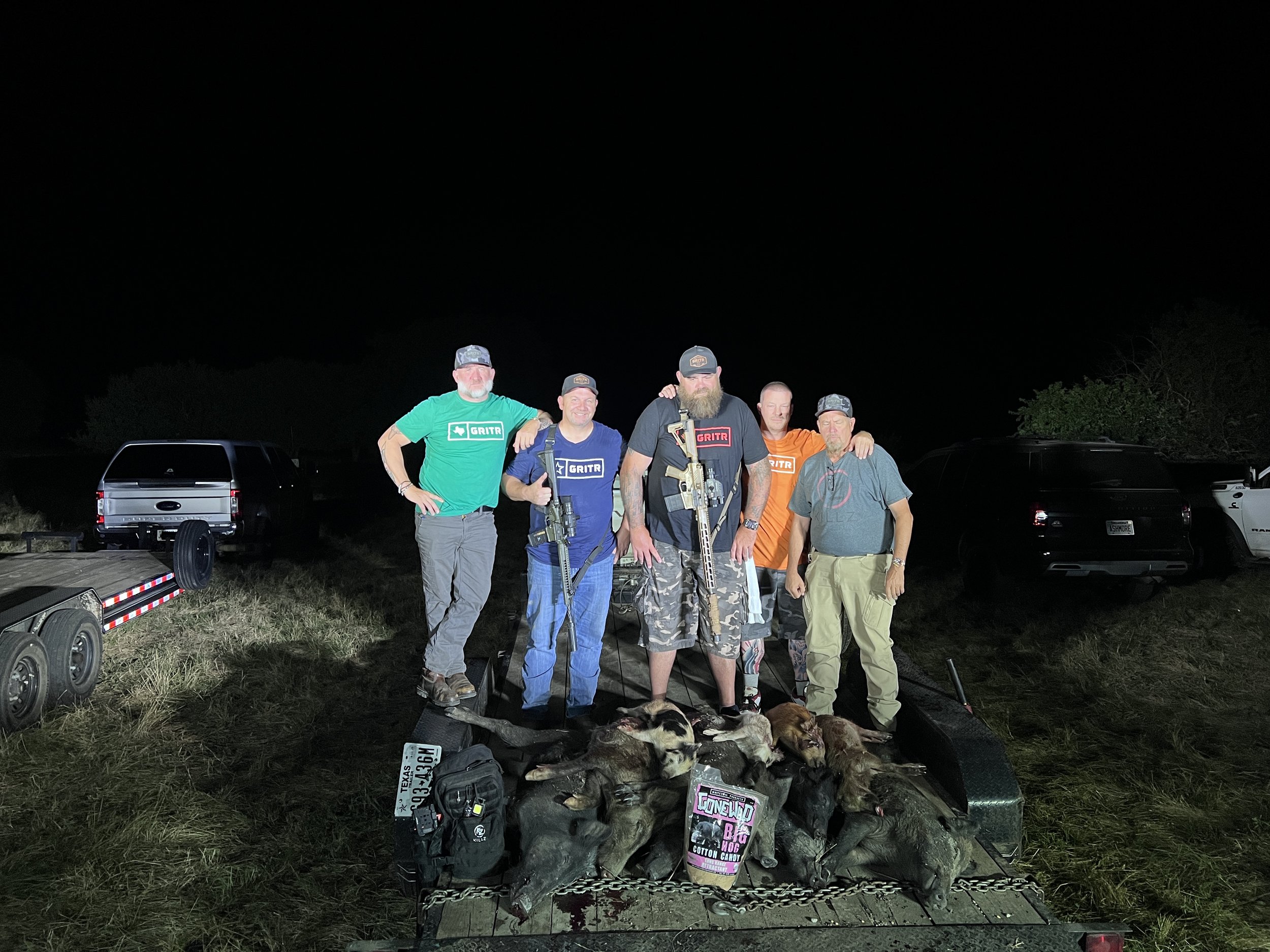A group of five men standing outdoors at night, posing for a photo, with a table of hunted animals and hunting gear in front of them.
