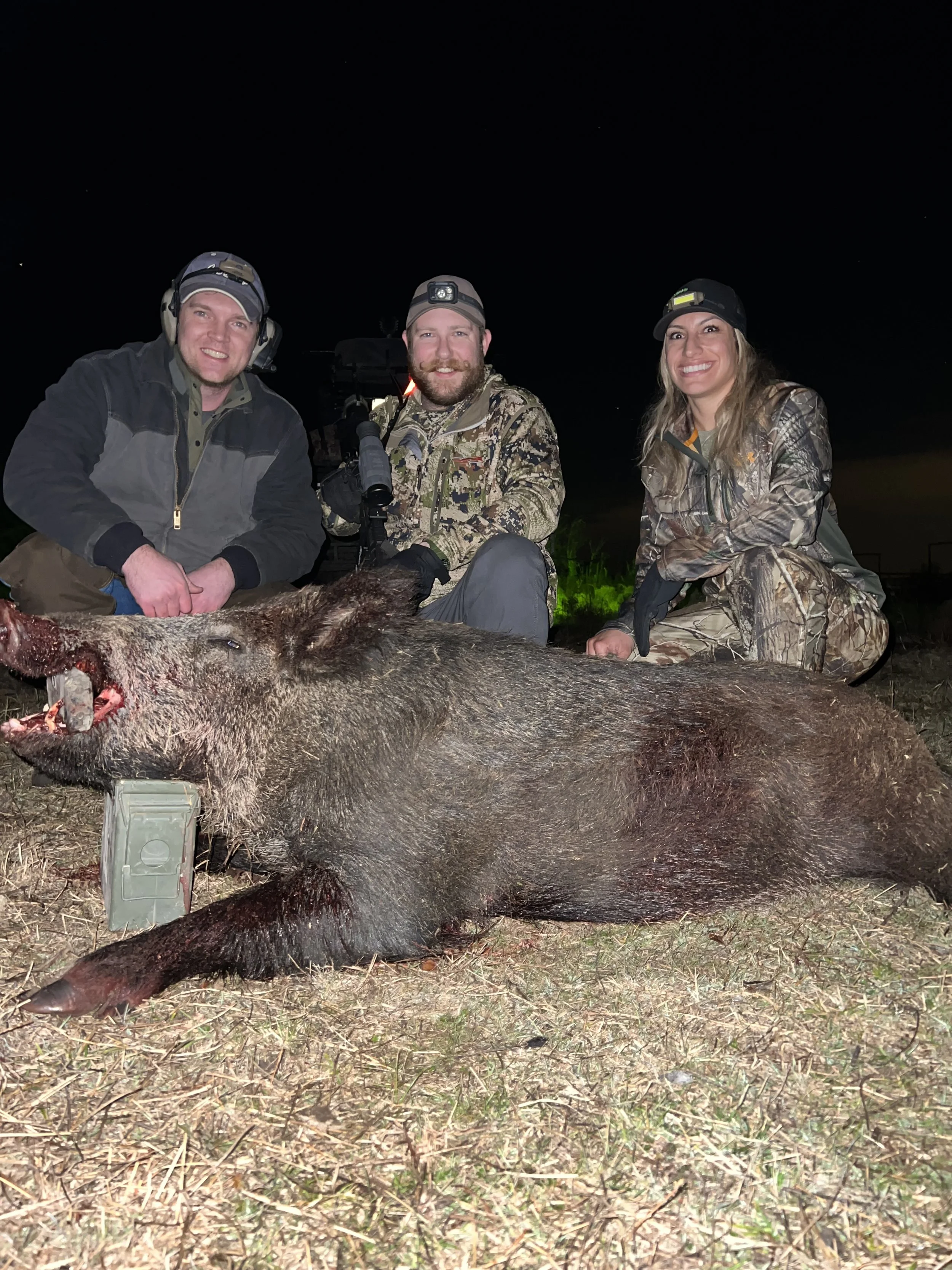 Three people in camouflage clothing and headlamps, smiling, kneeling behind a large, dead wild boar that is lying on the ground at night.