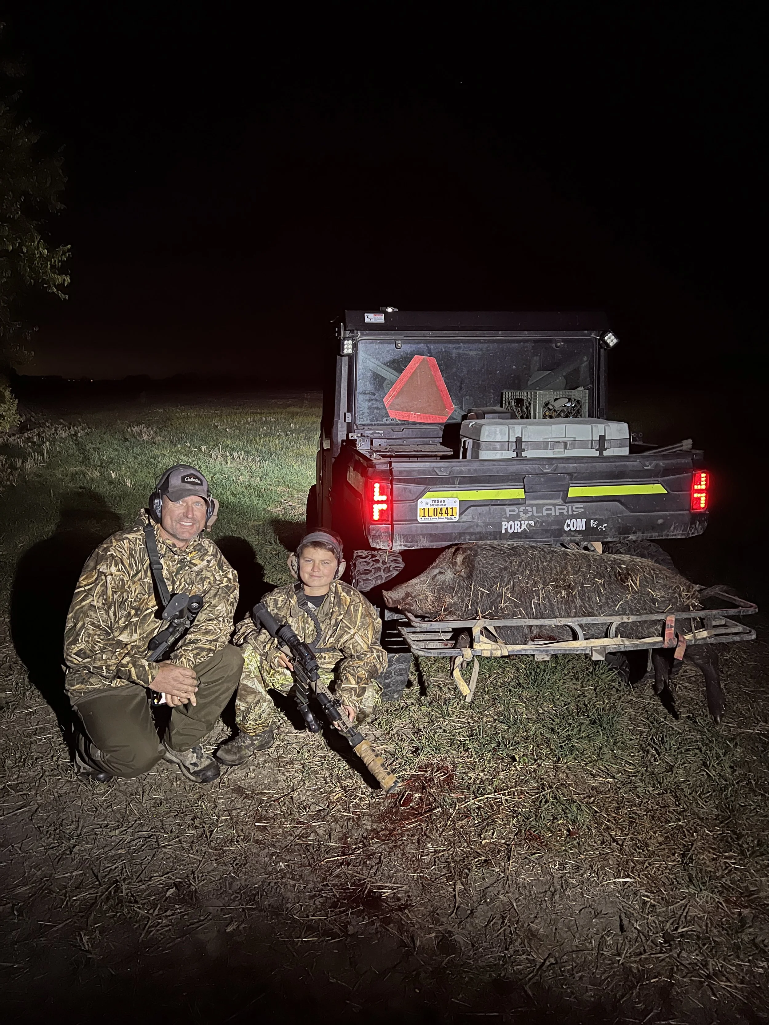 Nighttime outdoor scene with a man and a boy dressed in camouflage gear, kneeling on grass beside a trailer with a hog on it, in front of a black pickup truck with a red reflective triangle at night.