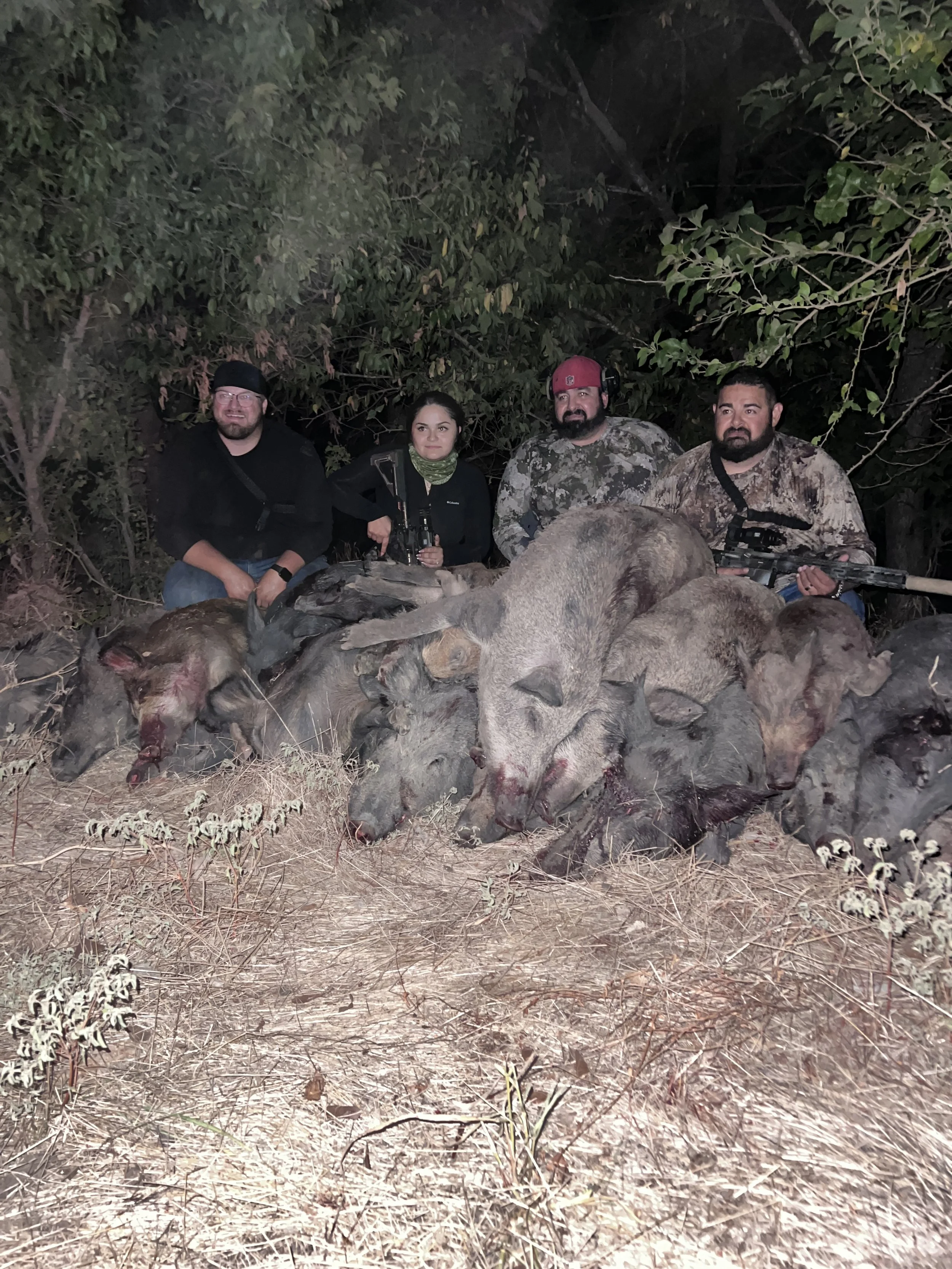 Group of four people in camouflage and dark clothing sitting behind a pile of dead wild animals, likely for hunting, at night in a wooded area.
