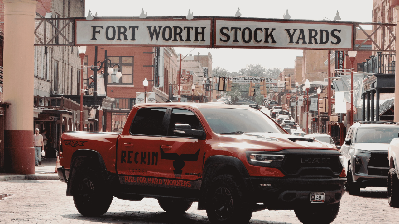 Red pickup truck with 'Reckin' and 'Fuel for Hard Workers' graphics parked under a sign that reads 'Fort Worth Stock Yards' in a historic downtown area.