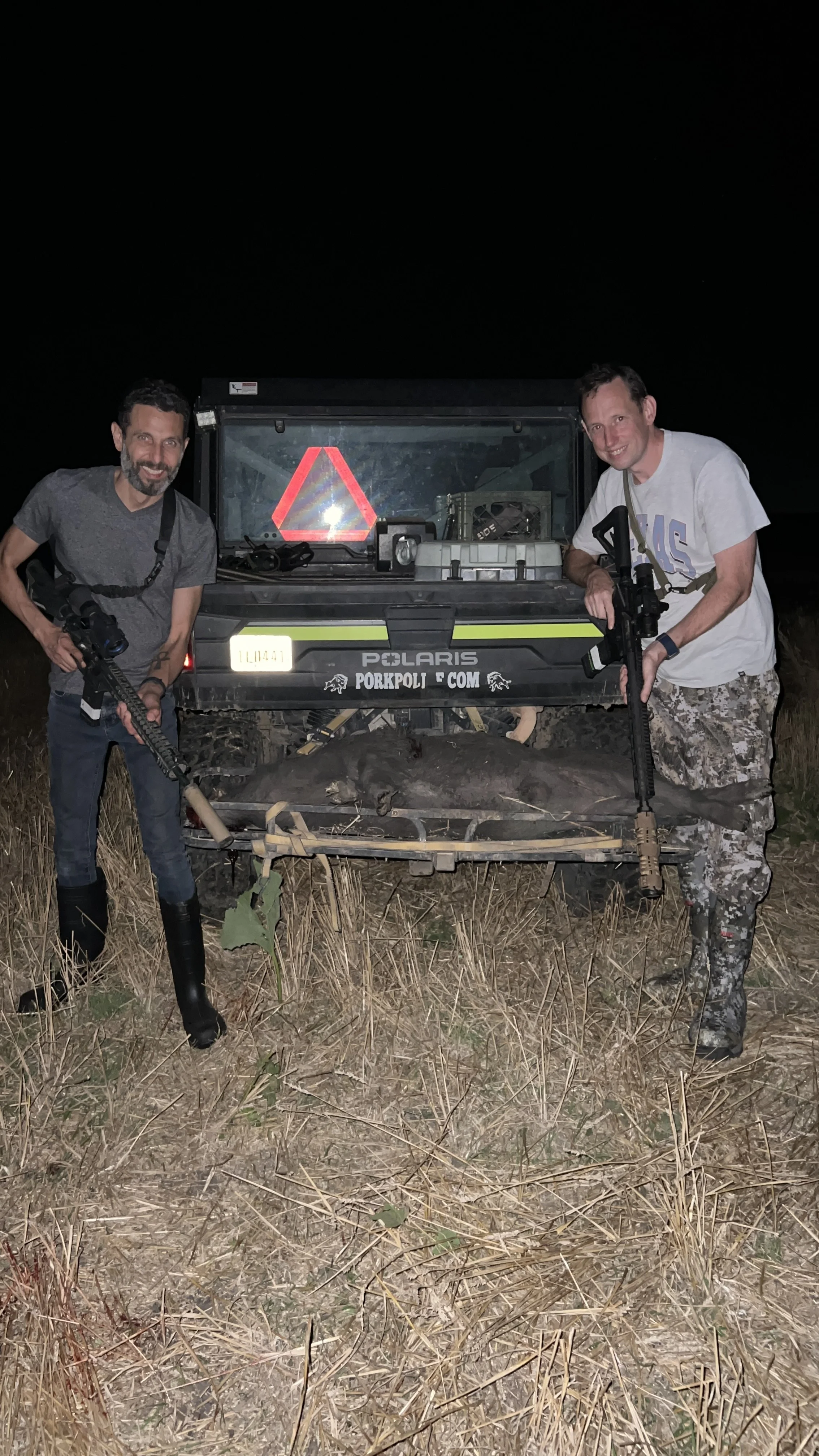 Two men standing outdoors at night, each holding a rifle, next to a Polaris off-road vehicle in a field of dry grass.
