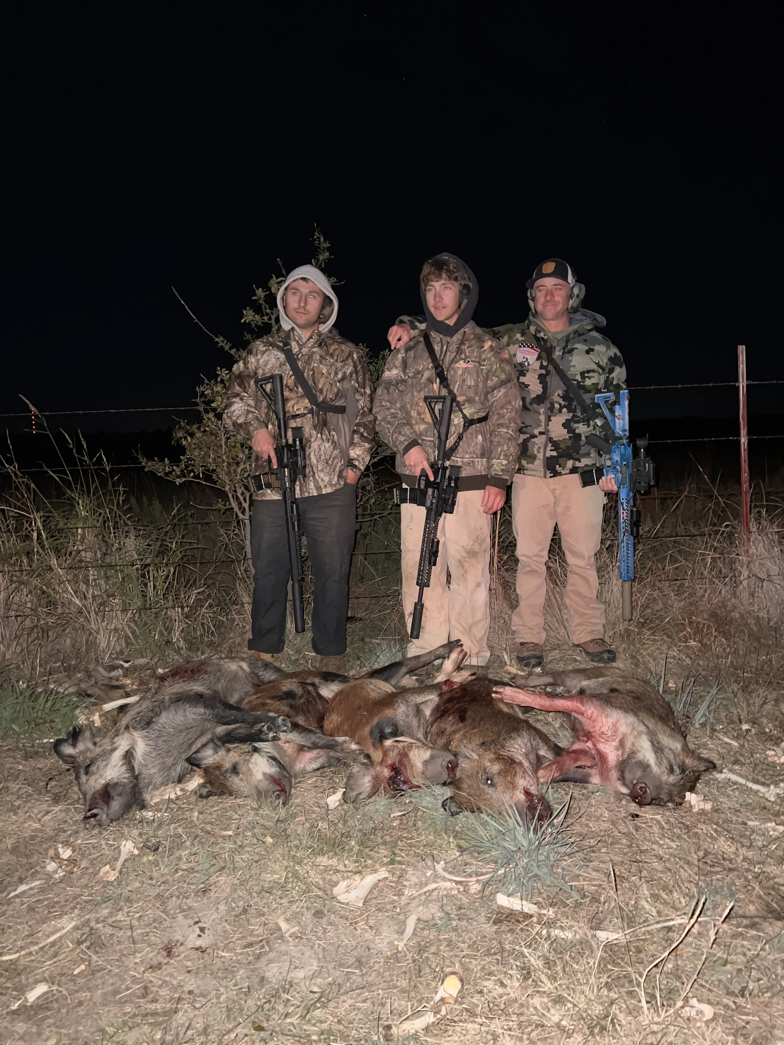 Three hunters standing outdoors at night with rifles, posing behind a row of dead animals including porcupines and wild boars.