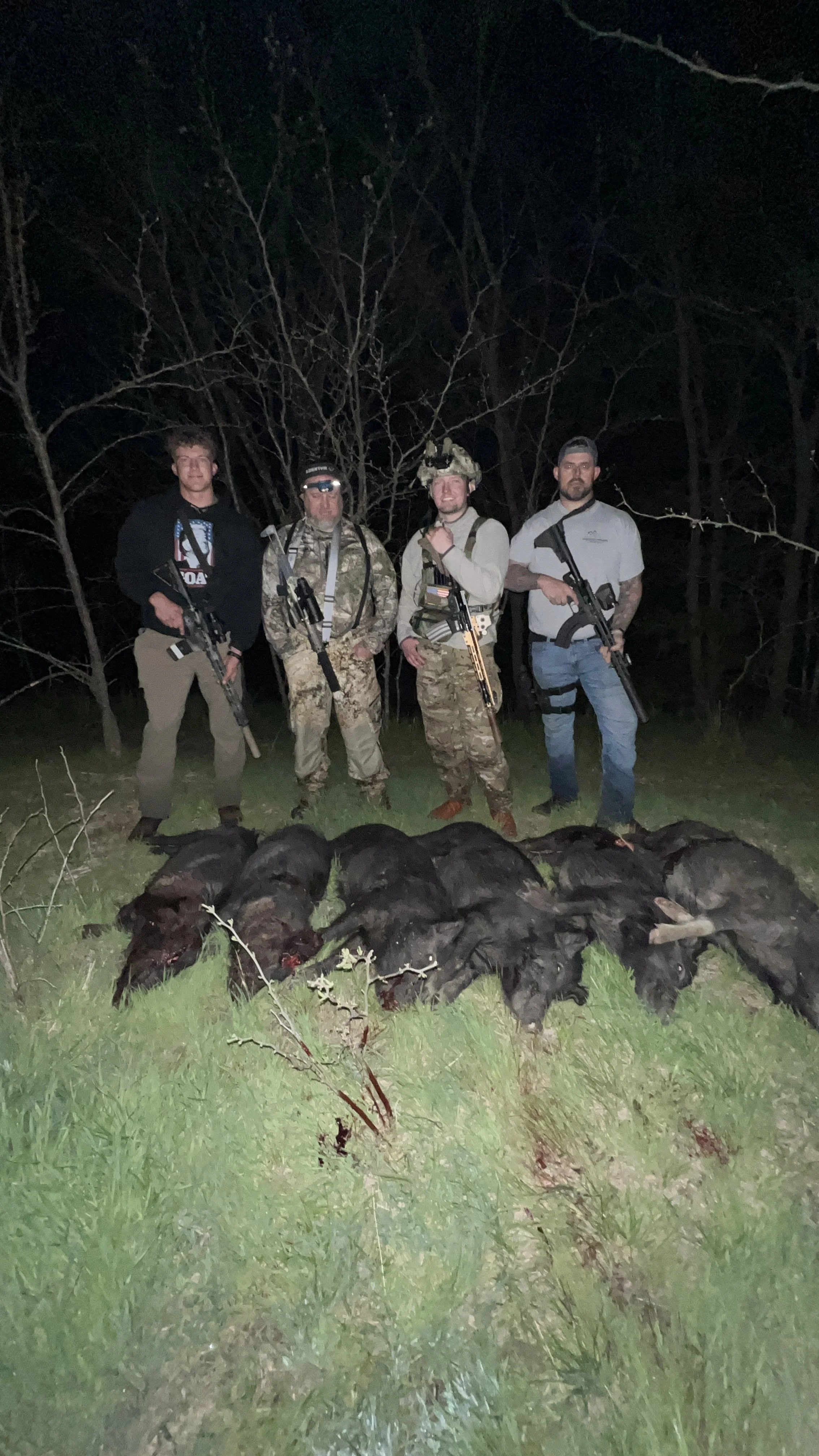 Four men standing outdoors at night behind a line of dead animals, possibly hunting game, with trees in the background and grass in the foreground.