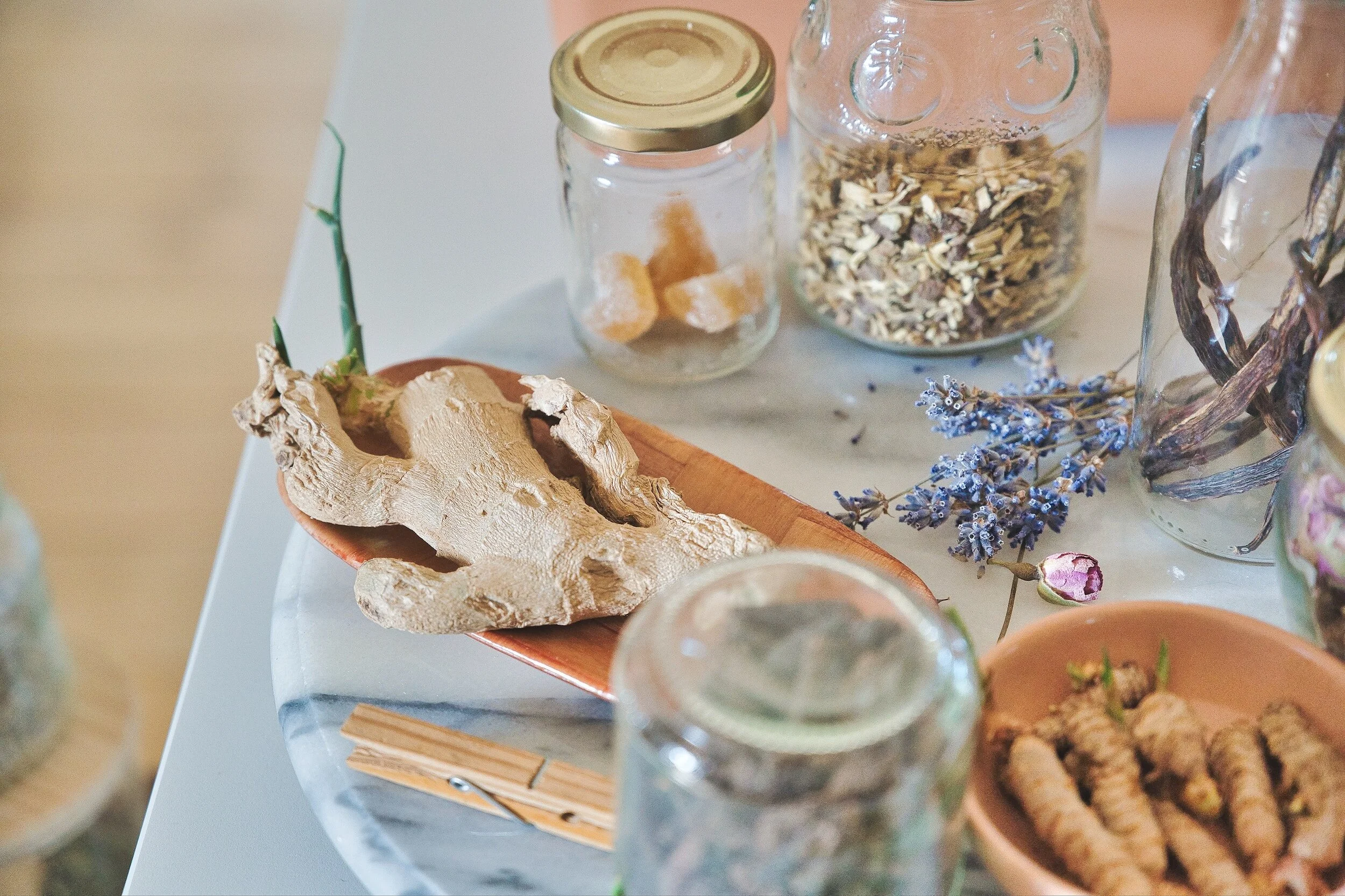 Assorted dried herbs and spices, including ginger root on a wooden dish, jars with dried ingredients, and lavender flowers on a marble surface.