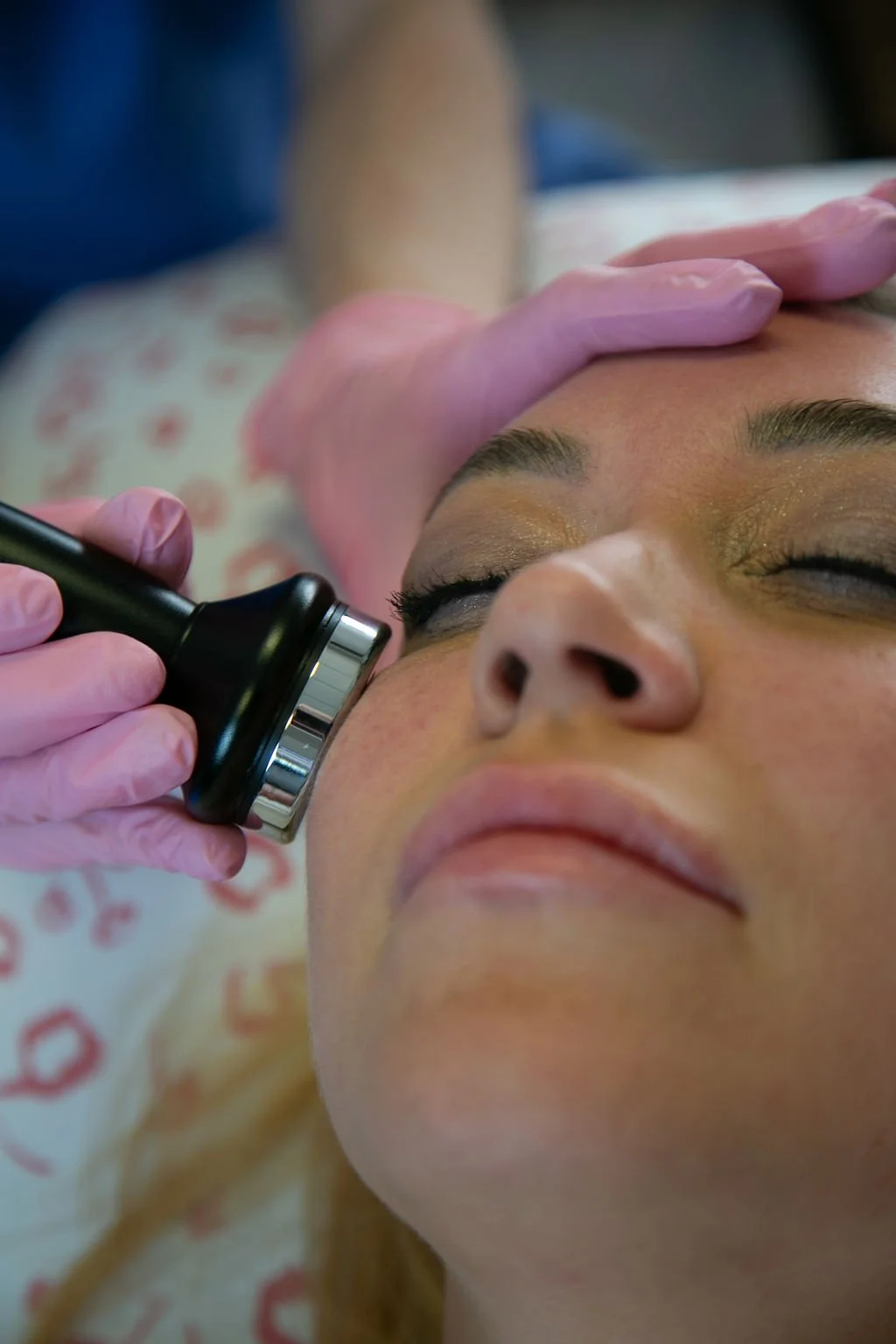 Person receiving a facial treatment with a black device, lying down with closed eyes, while a professional applies the treatment and supports their head.
