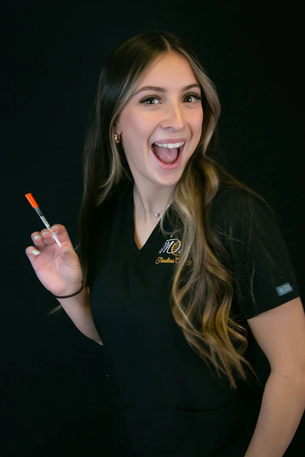 A young woman with long, wavy hair holds a syringe in her right hand and smiles widely against a black background, wearing a black shirt with embroidery.