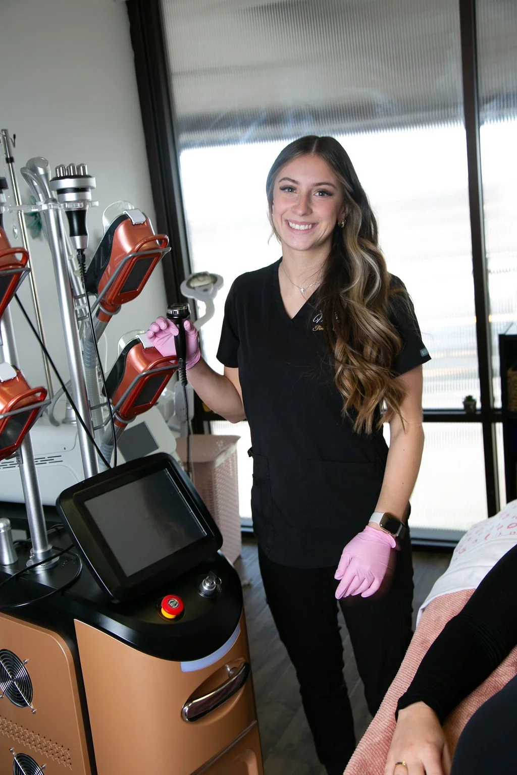 A smiling woman in medical scrubs and pink gloves standing next to laser equipment in a clinic.