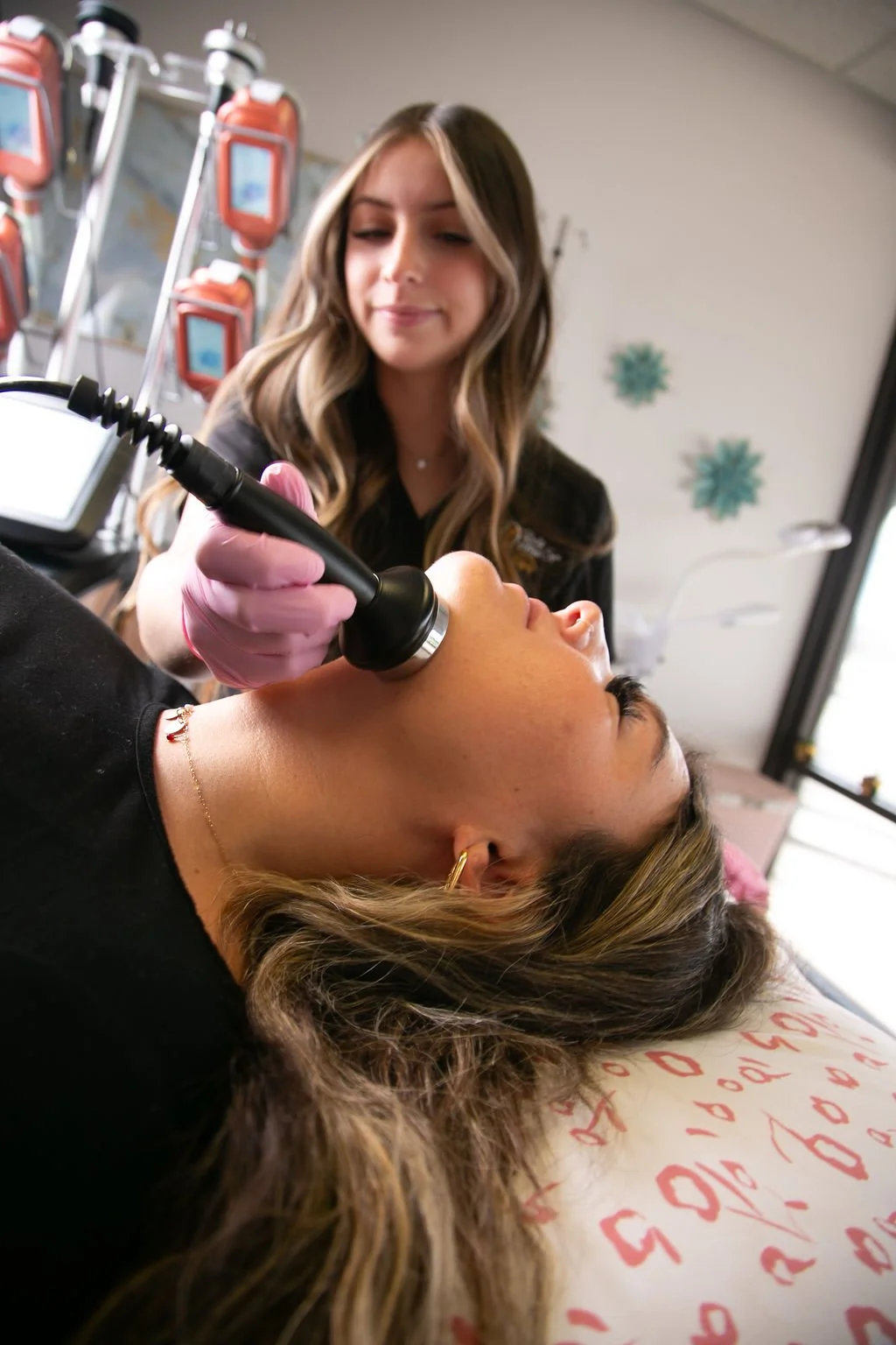 A woman lying down with her eyes closed as a technician performs a skin treatment with a handheld device. The technician is wearing pink gloves and smiling, in a clinical setting with medical equipment and decorative snowflakes on the wall.
