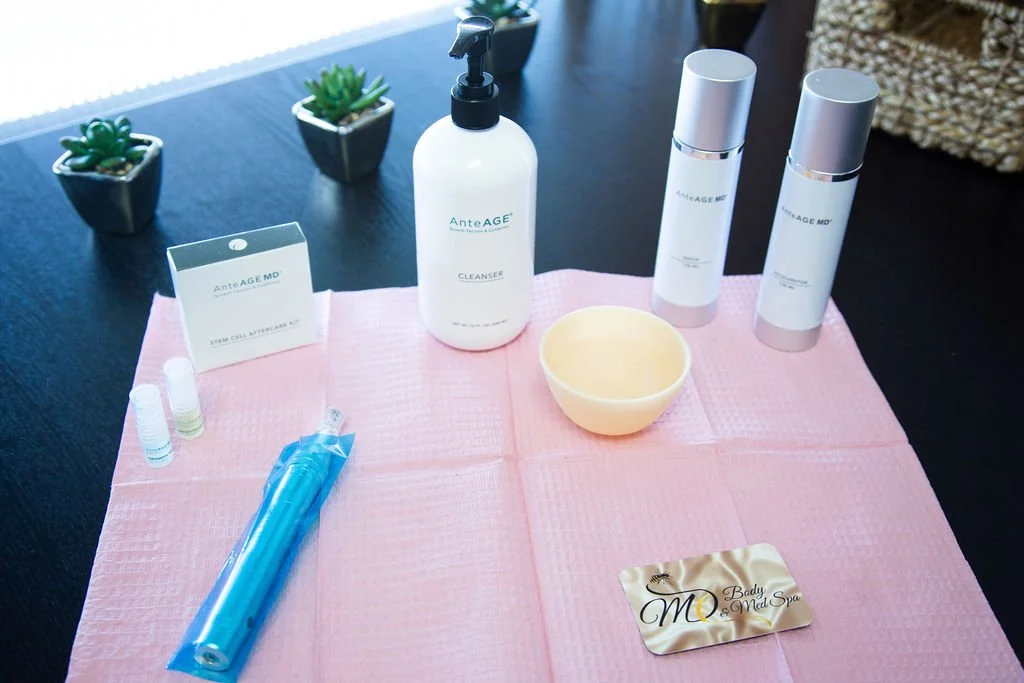 Skincare products, including cleansers and serums, a bowl, and small vials on a pink cloth, with potted succulents in the background.