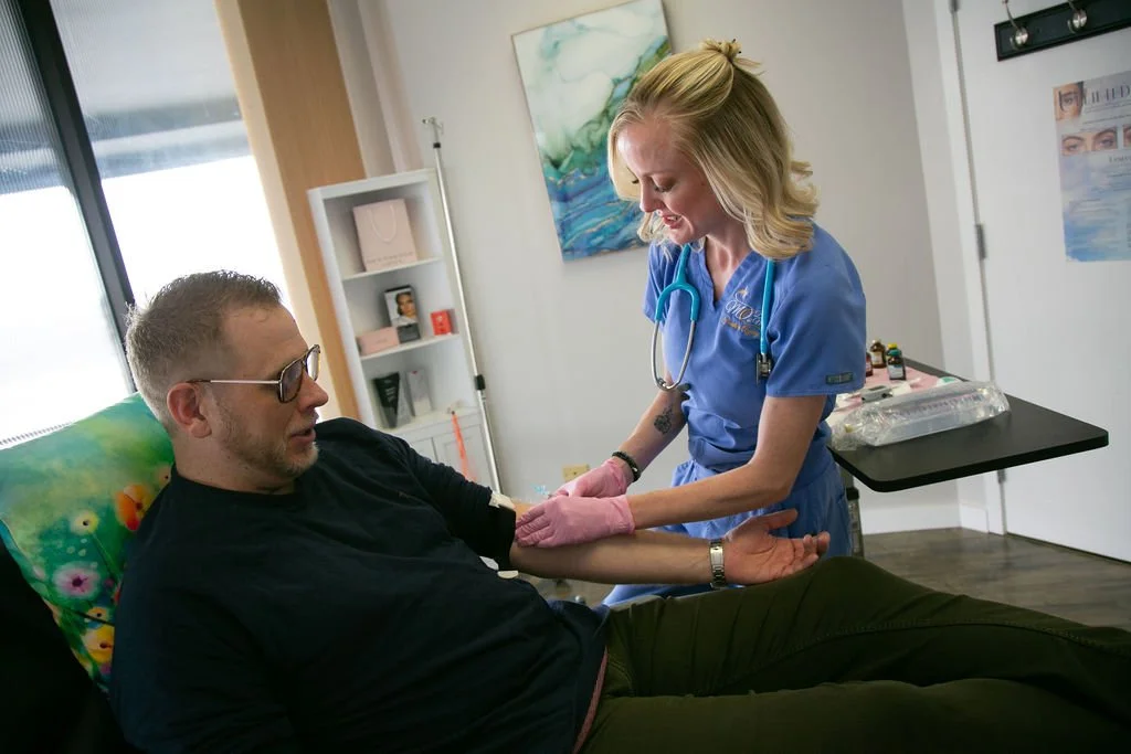 A healthcare worker, wearing pink gloves and a blue uniform, is administering a medical test or treatment to a man sitting in a chair. The man is wearing glasses and a black shirt, and is sitting in a room with medical supplies and artwork visible in the background.