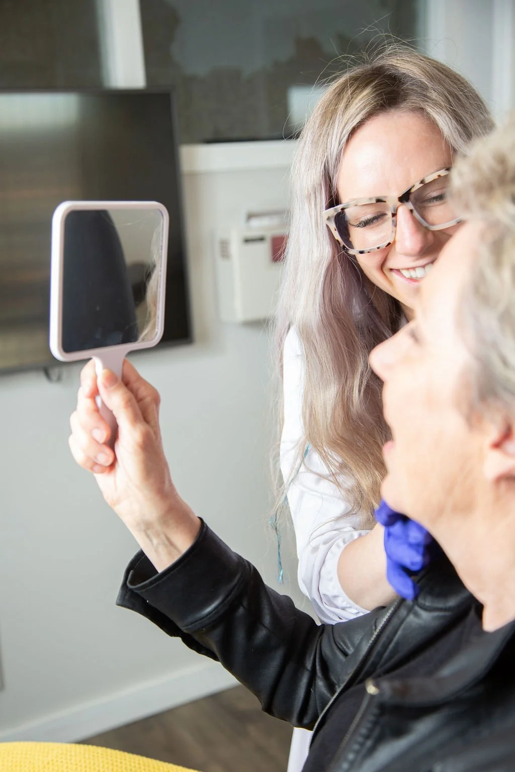 A woman with glasses smiling and holding a mirror, close to an elderly woman, in a room with neutral-colored walls.