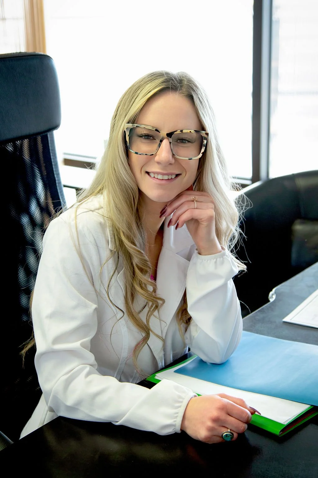 A young woman with long blonde hair and glasses, smiling and resting her chin on her hand, sitting at a desk with folders and papers in a bright office setting.