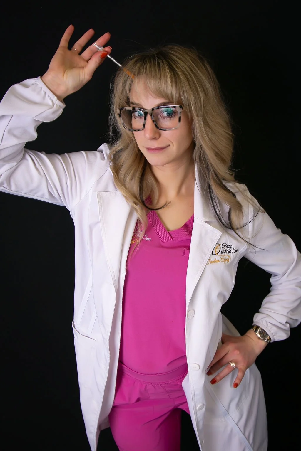 A woman in pink medical scrubs and a white medical coat, wearing glasses, holding a syringe, posing against a black background.
