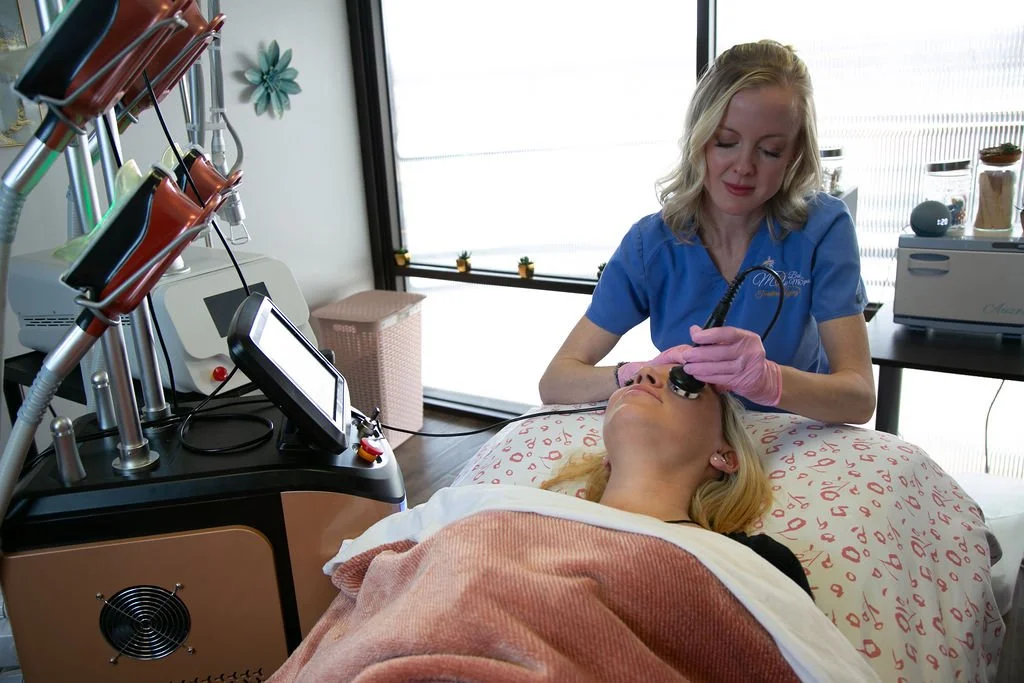 A woman receives a cosmetic facial treatment with a small device on her face in a professional clinic setting, with a technician wearing pink gloves and a blue uniform performing the procedure.