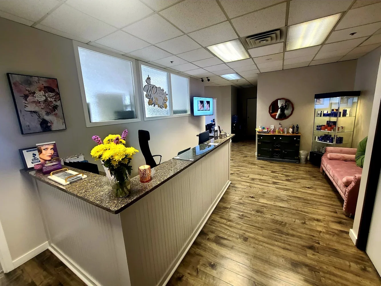 Reception area featuring a front desk with a flower arrangement, seating area with a pink velvet couch, a display case with products, and a wall-mounted TV.