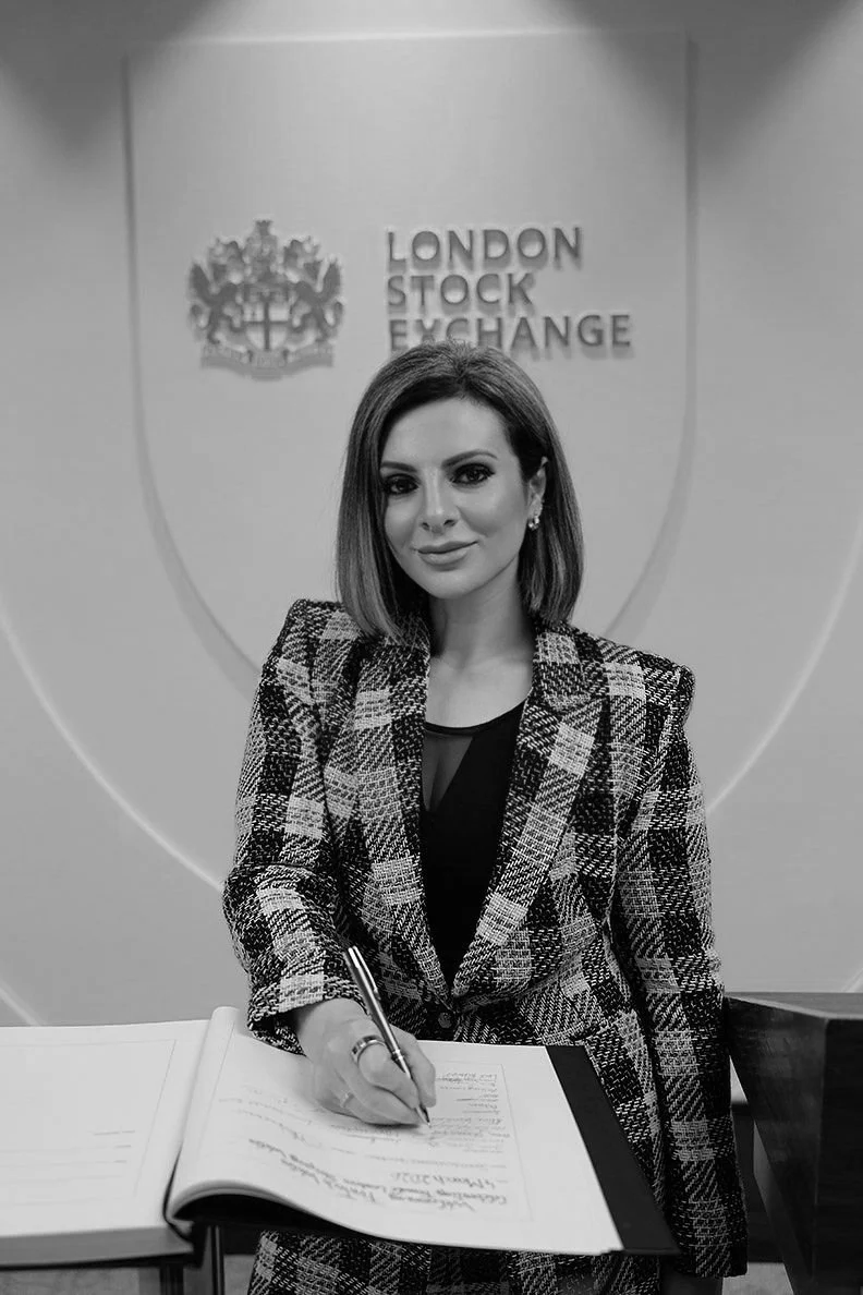 Lucy Cohen wearing a plaid blazer, sitting at a table and signing a document. In the background, there is a sign that reads 'LONDON STOCK EXCHANGE' with a crest above it.