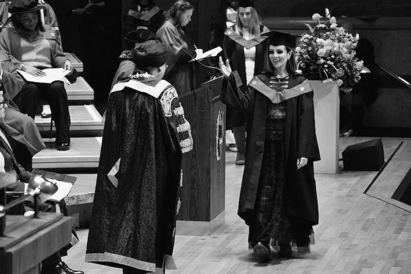 A graduation ceremony with Lucy Cohen in cap and gown walking across the stage to receive her Masters from a university official.