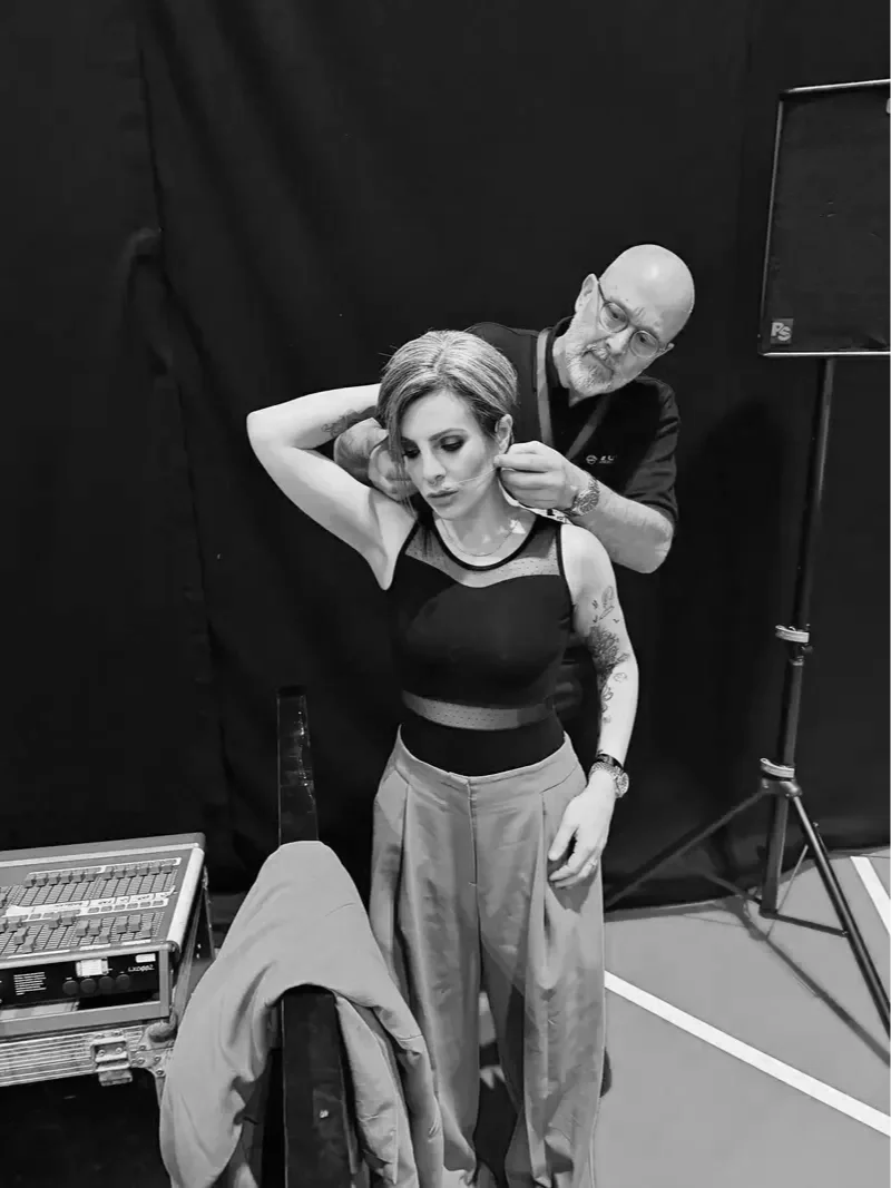 Lucy Cohen  with styled hair and tattoos on her arm being assisted by a man, possibly a stylist, in a professional setting with studio equipment visible.
