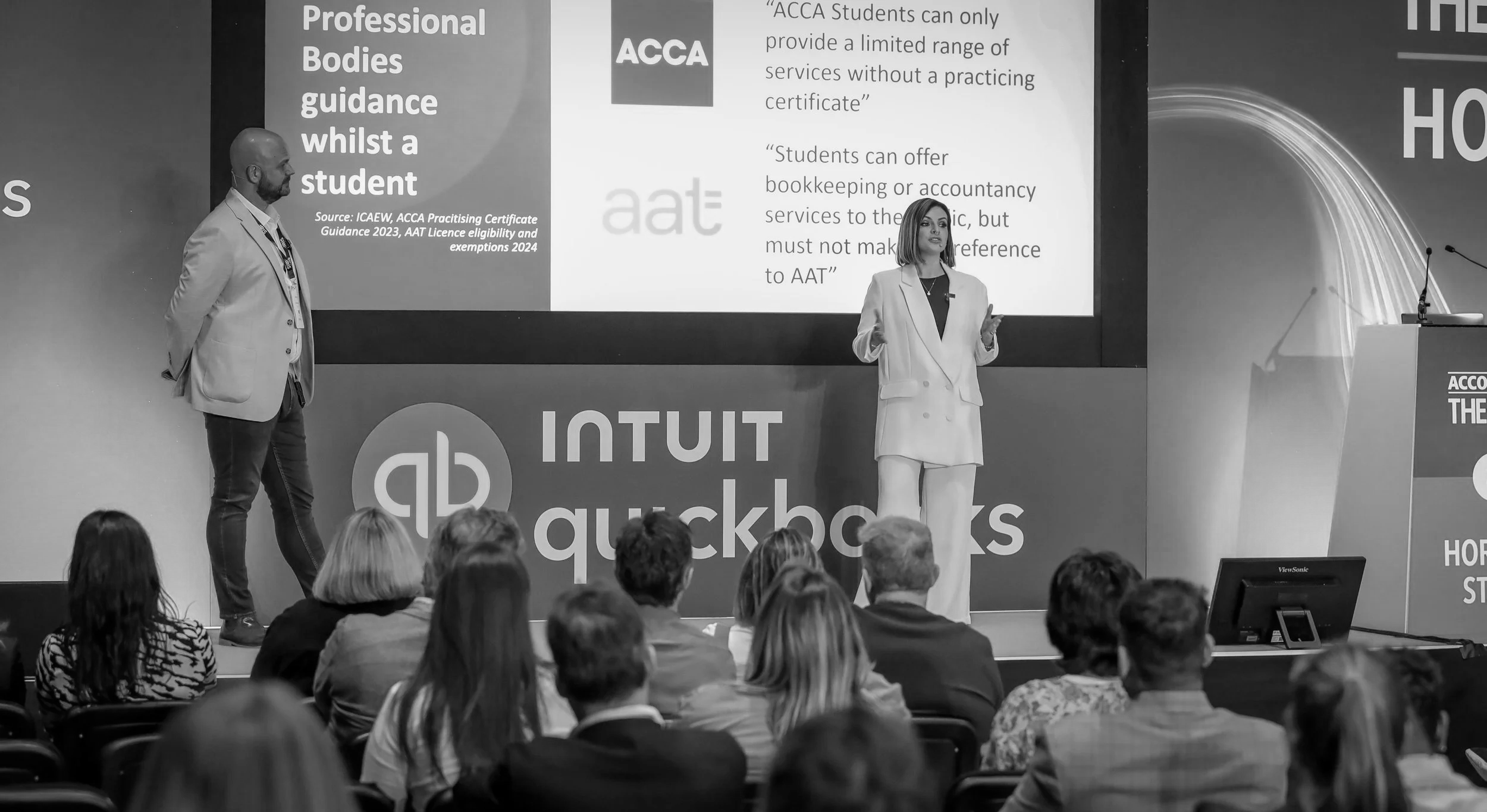 Lucy Cohen and another presenter on stage at an event, speaking to an audience. The background shows a large screen with text about professional bodies guidance and ACCA. The audience is seated and listening attentively.