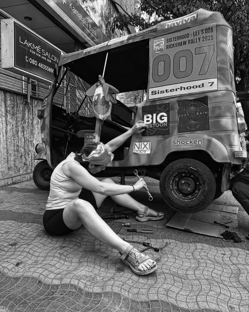 Lucy Cohen repairing a black auto rickshaw on the sidewalk, sitting on the ground with tools around her.