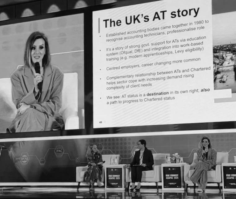 A panel discussion at a conference with a large screen displaying a presentation titled 'The UK's AT story.' The screen has bullet points about the history and development of accounting technicians in the UK. Three people are seated on stage, two women and one man, engaged in conversation.