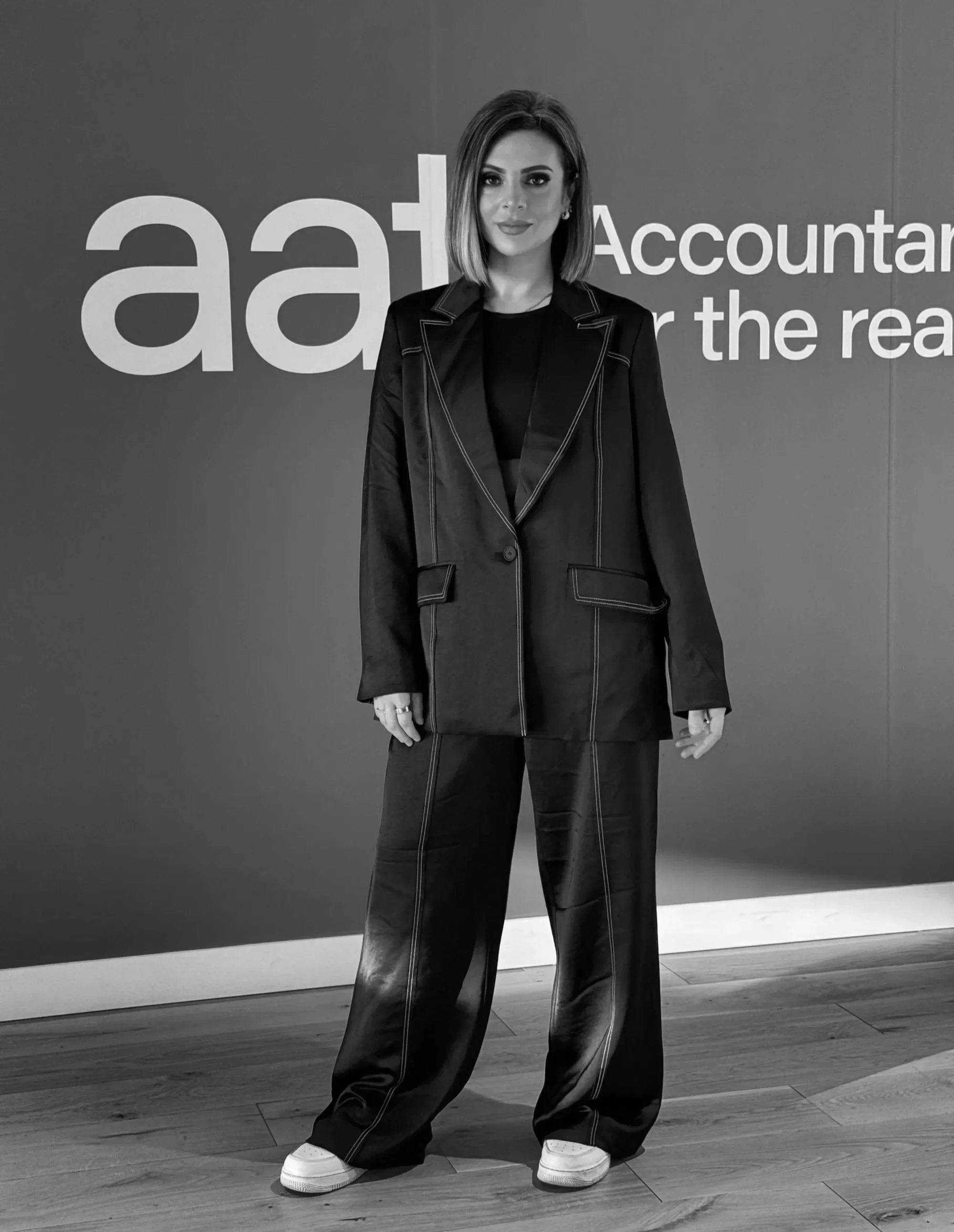 Lucy Cohen in a blazer and wide-leg pants standing in front of a wall with the text 'accountant' and 'AAT. She has short hair and is wearing sneakers.