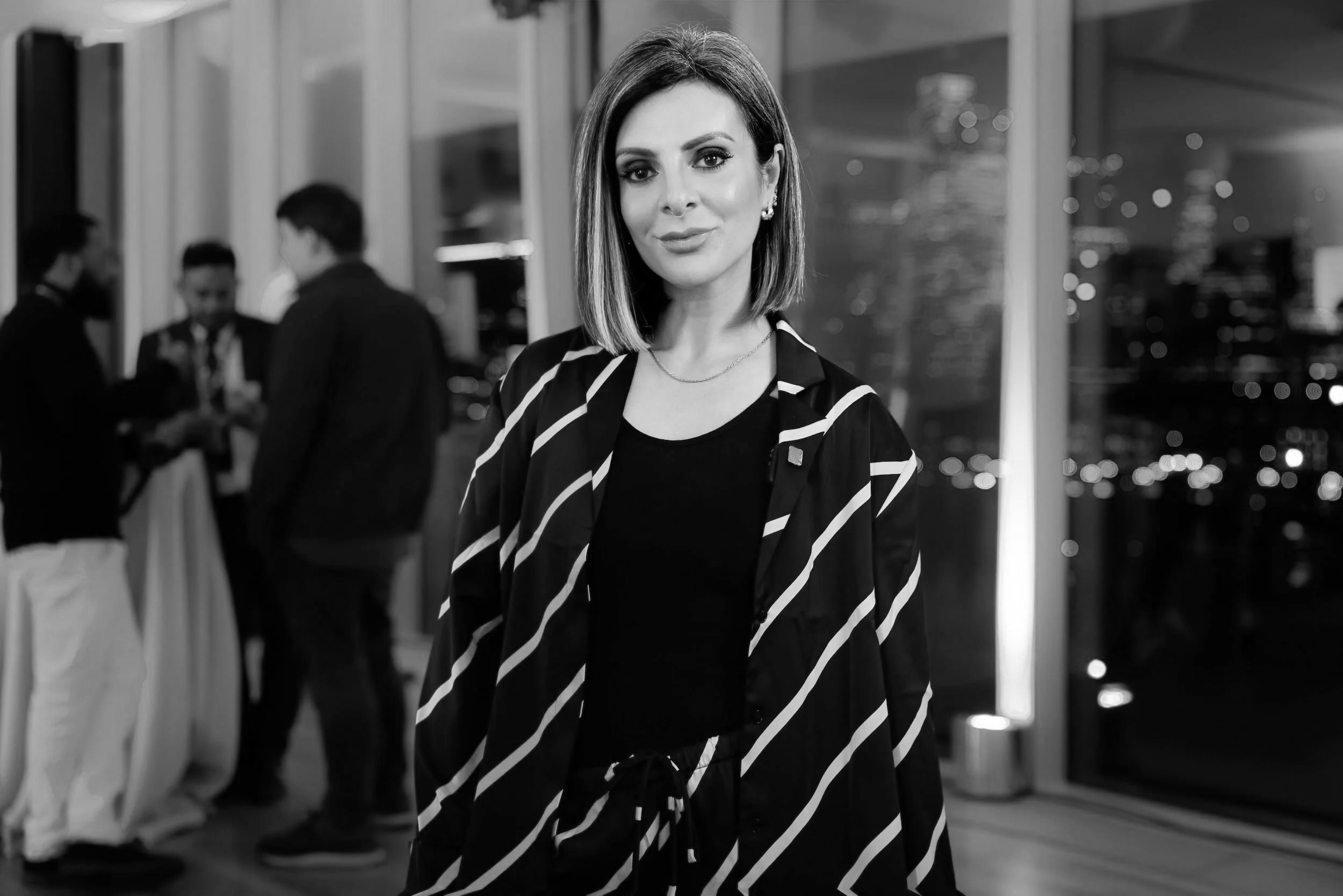 Lucy Cohen with shoulder-length hair and earrings, wearing a striped blazer, looking at the camera at an indoor event with people in the background.