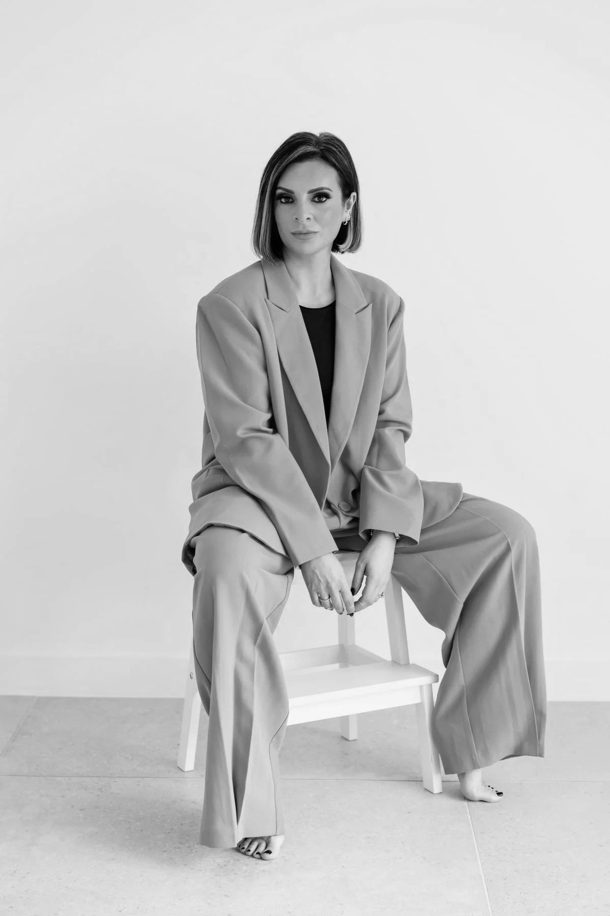 Lucy Cohen sitting on a chair against a plain background, wearing a suit with a blazer and wide-leg trousers, with one foot on the floor and the other resting on the chair's rung. She has short hair and is looking directly at the camera.