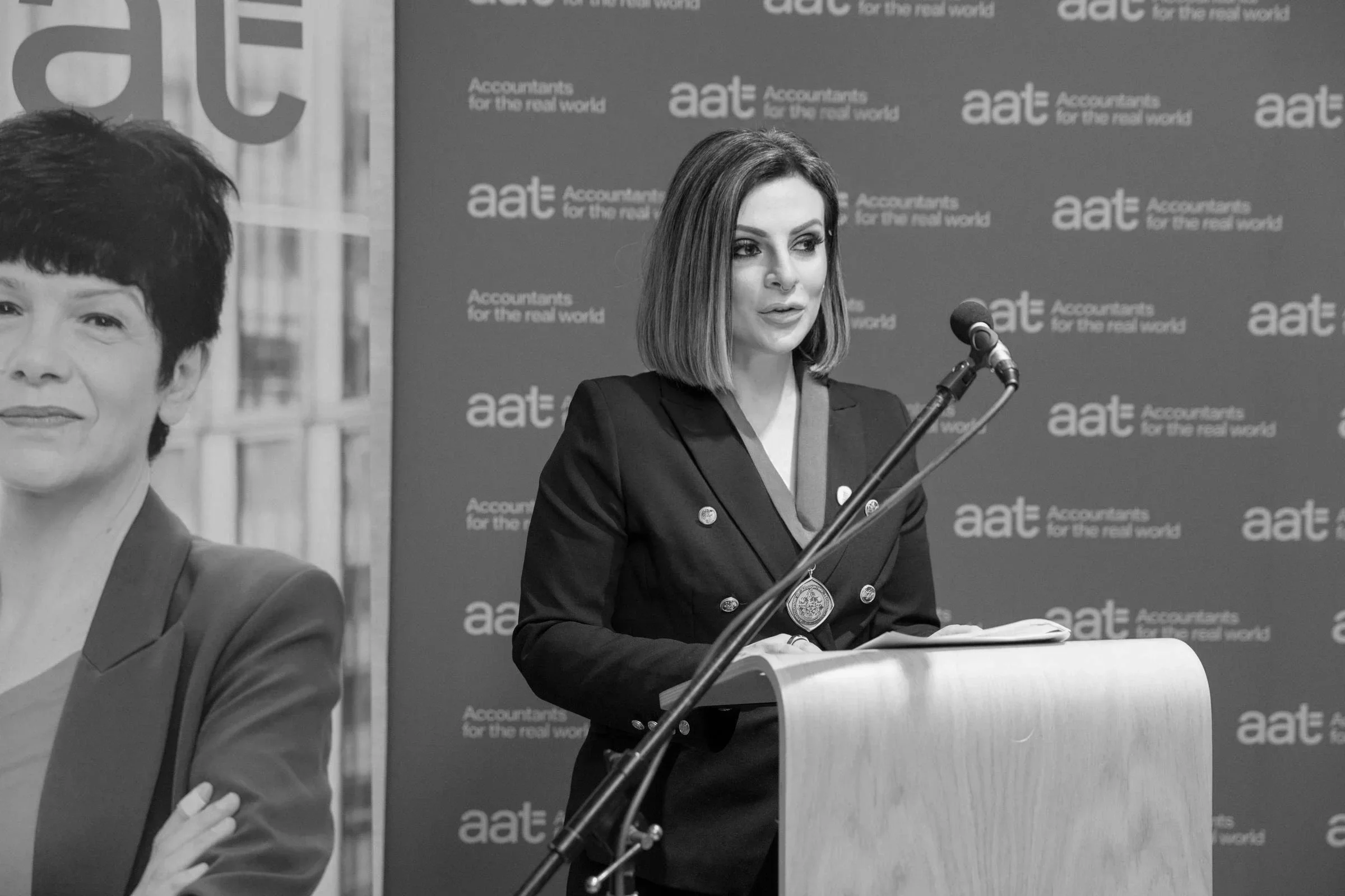 Lucy Cohen speaking at a microphone during a press conference or event, with a backdrop that reads 'aat' and 'Accountants for the real world.'