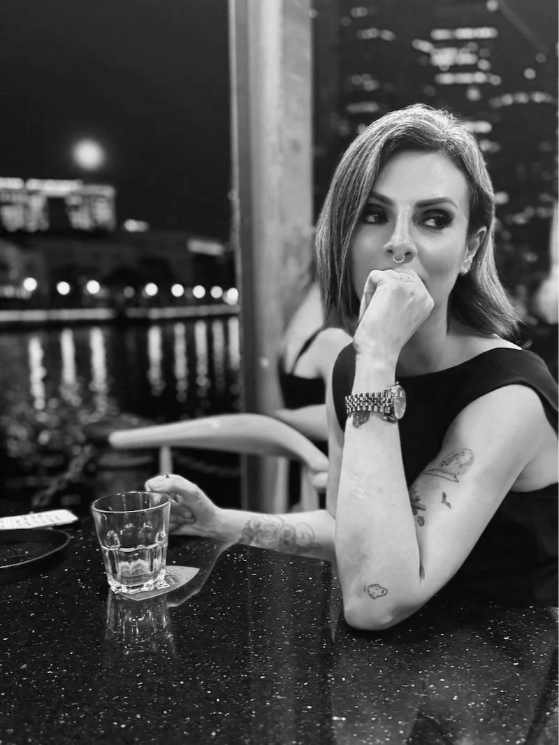 Lucy Cohen sitting at a table with a glass, in a dark, urban setting near a body of water at night, with city lights and buildings in the background.