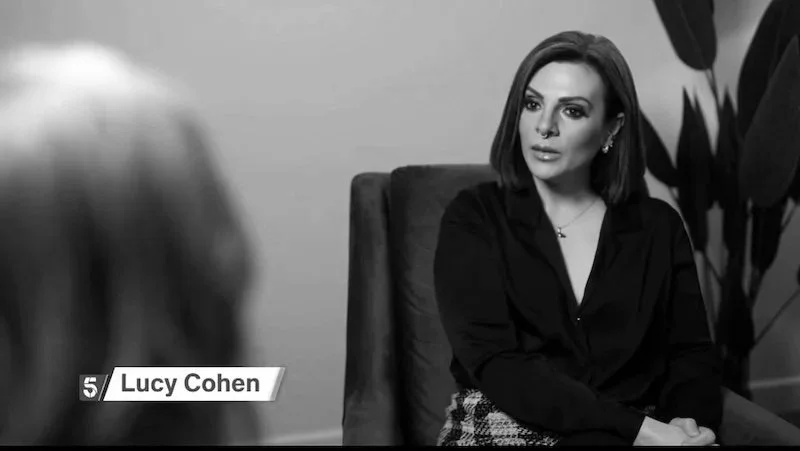 A woman with shoulder-length dark hair wearing a black top sitting in an office chair during an interview or conversation. A blurred figure with light-colored hair is in the foreground. Text overlay reads "Lucy Cohen" with a number '5' icon.