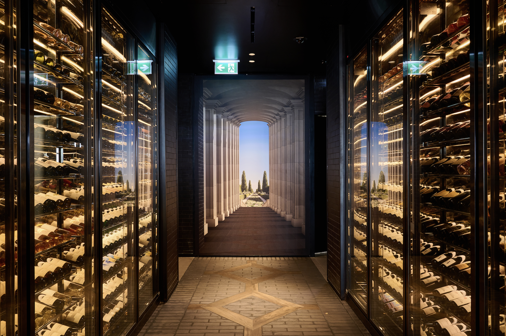 A Wine cellar hallway containing a variety of red and white wine being displayed behind lit glass displays, featuring a large painting at the end of the corridor depicting white pillars leading towards a luscious garden.