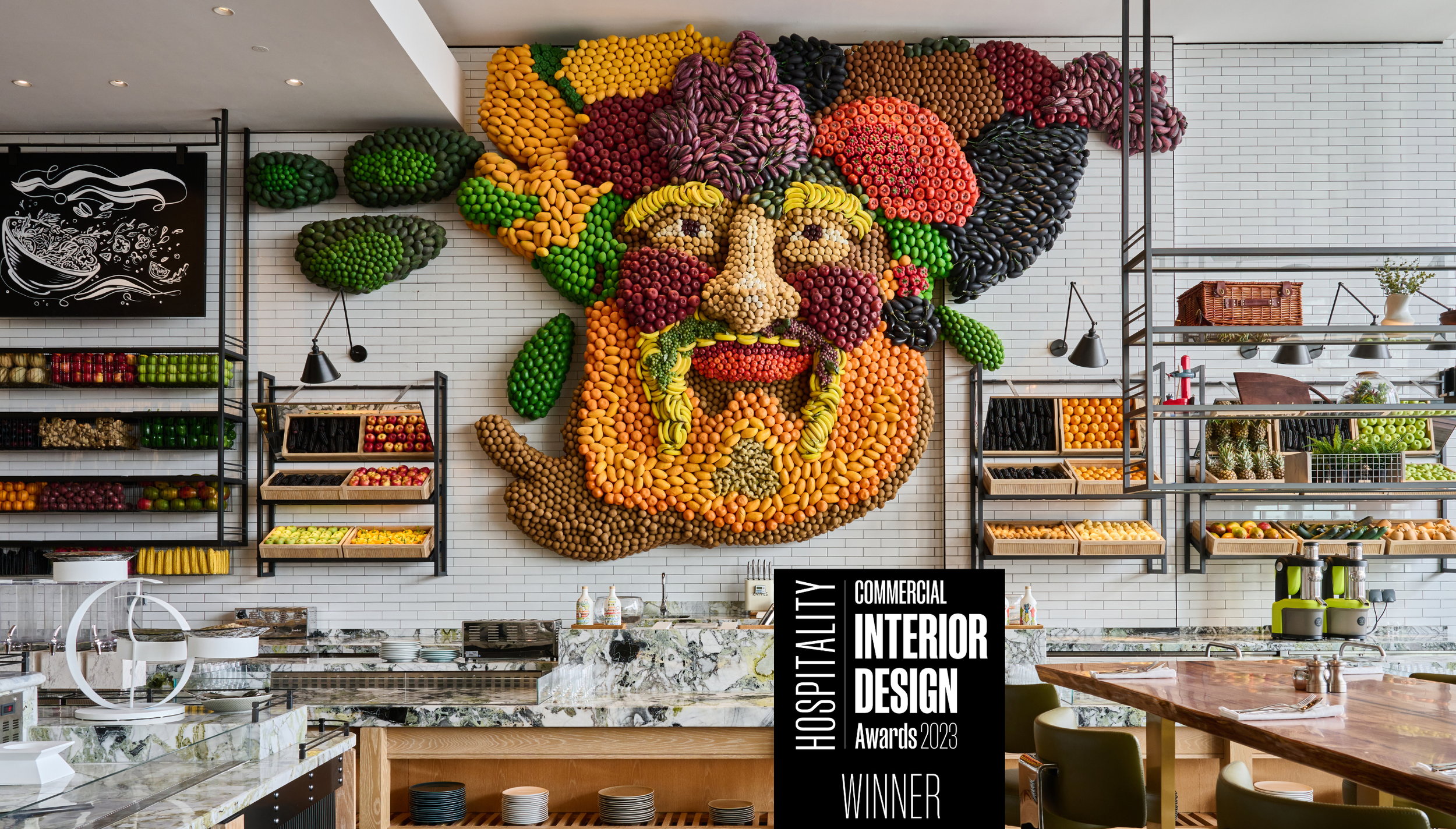 An interior space with a large wall art installation made of fruits forming a portrait of a man with a beard and mustache. The space has shelves of fruits, a marble counter, and a sign indicating it is a winner of a hospitality interior design award.
