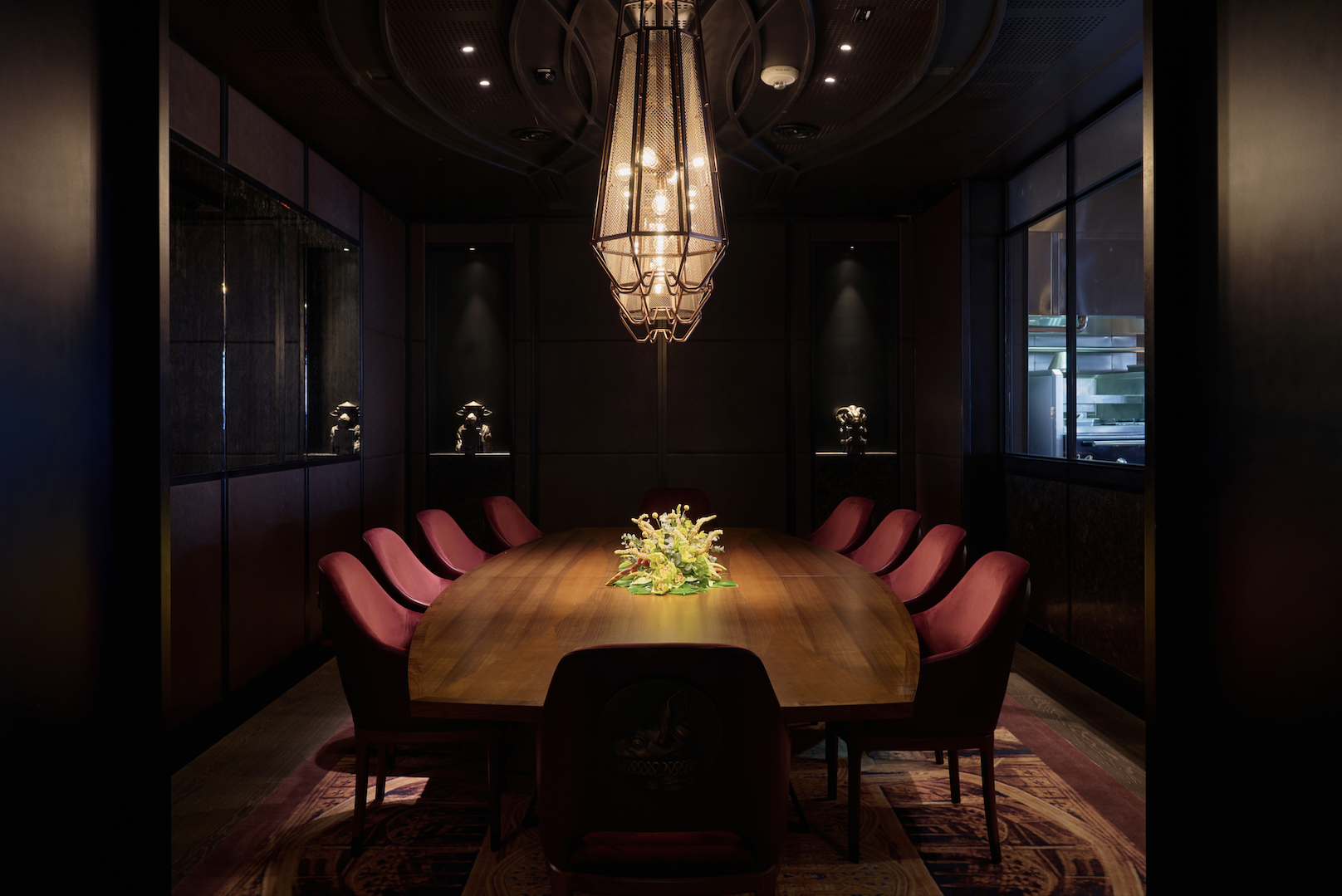 A dinner table in a dimly lit room with a lavish floral centrepiece and red chairs and a window into the kitchen, the room is decorated with historically inspired intricate metallic sculptures of different anthropomorphic animals.