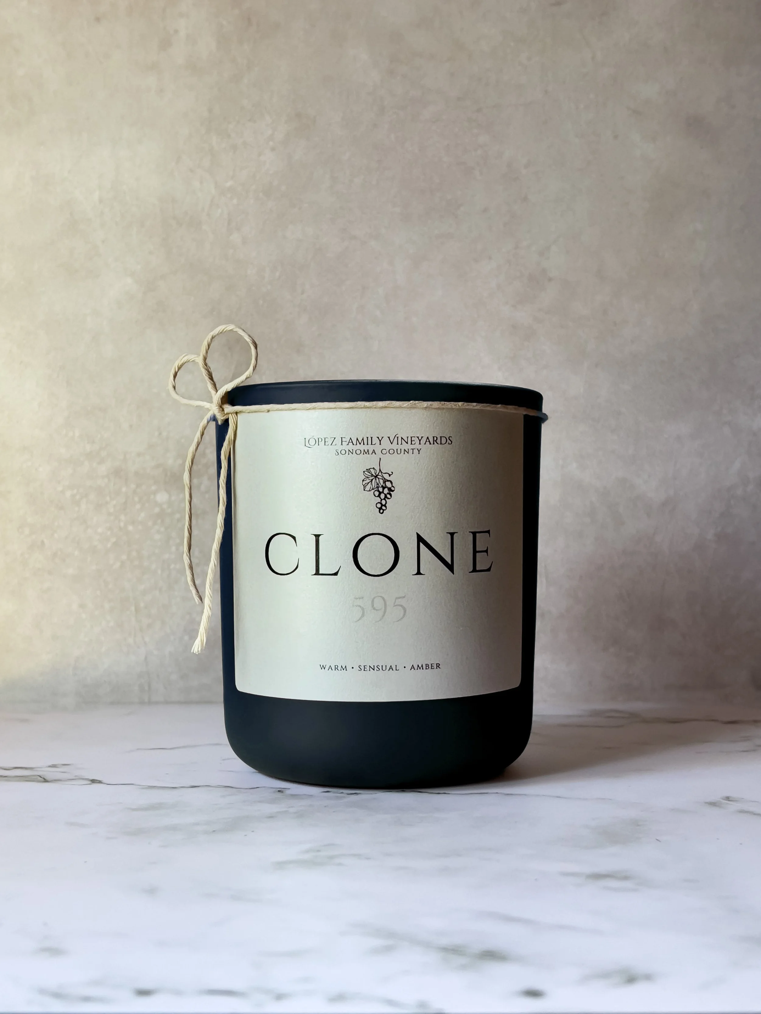 A black candle jar with a cream label reading 'Clone 595' from Lopez Family Vineyards, Sonoma County, with a decorative tied string left on the side, on a white marble surface against a gray textured background.