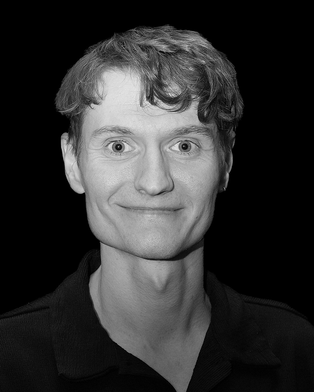 IMPROV COMEDY GBG Göteborg impro En man med brunt hår och blå denimskjorta står på scen och visar något med båda händerna, med en mörk bakgrund.