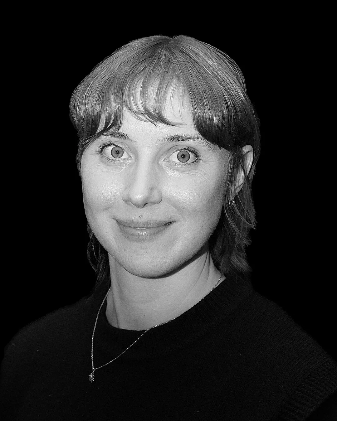 IMPROV COMEDY GBG Göteborg impro En ung kvinna med kort hår, vit skjorta och svarta byxor, som står på scen med en mikrofon. Hon tittar ner och håller i sina händer.