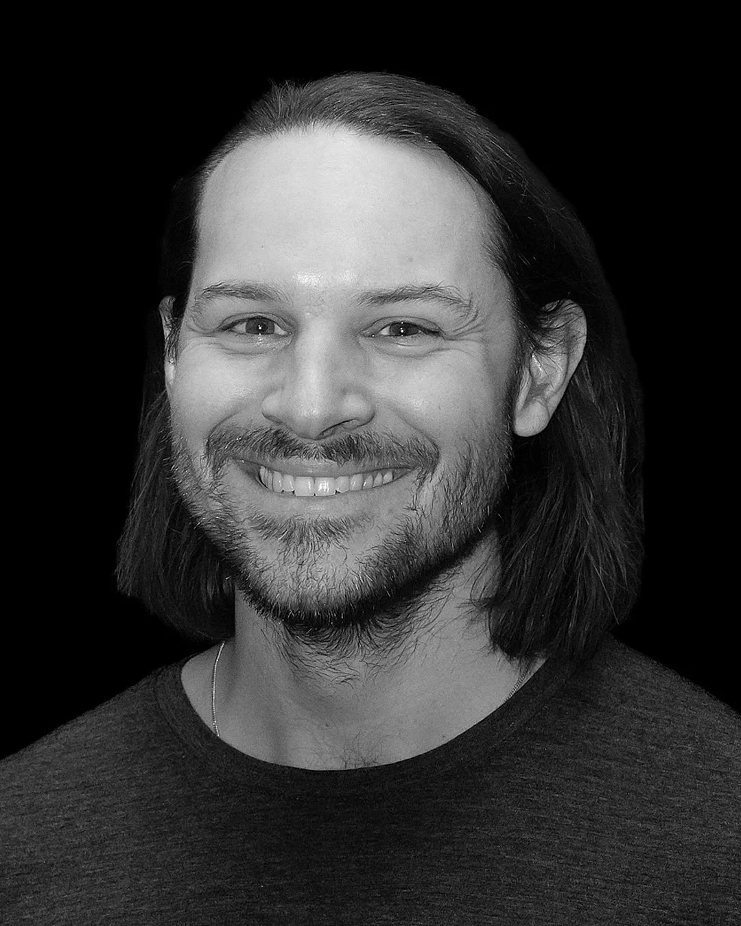 IMPROV COMEDY GBG Göteborg impro En man med svart hår, lätt skägg och vit t-shirt, tittar åt sidan i ett mörkt rum.