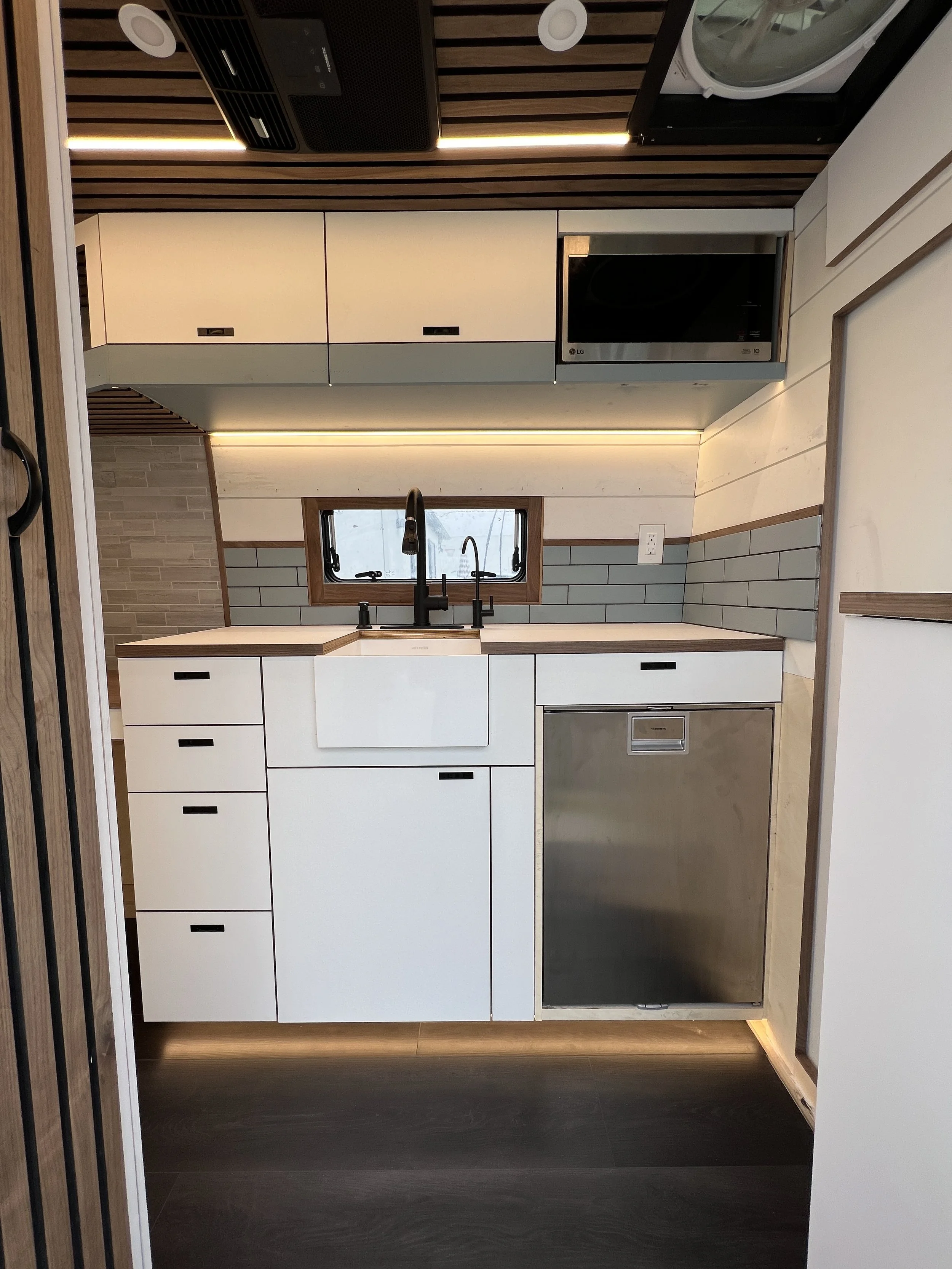 Small modern kitchen with white cabinets, a black sink, black faucet, gray subway tile backsplash, built-in microwave, and wooden accents, including a window above the sink and a wooden ceiling panel.