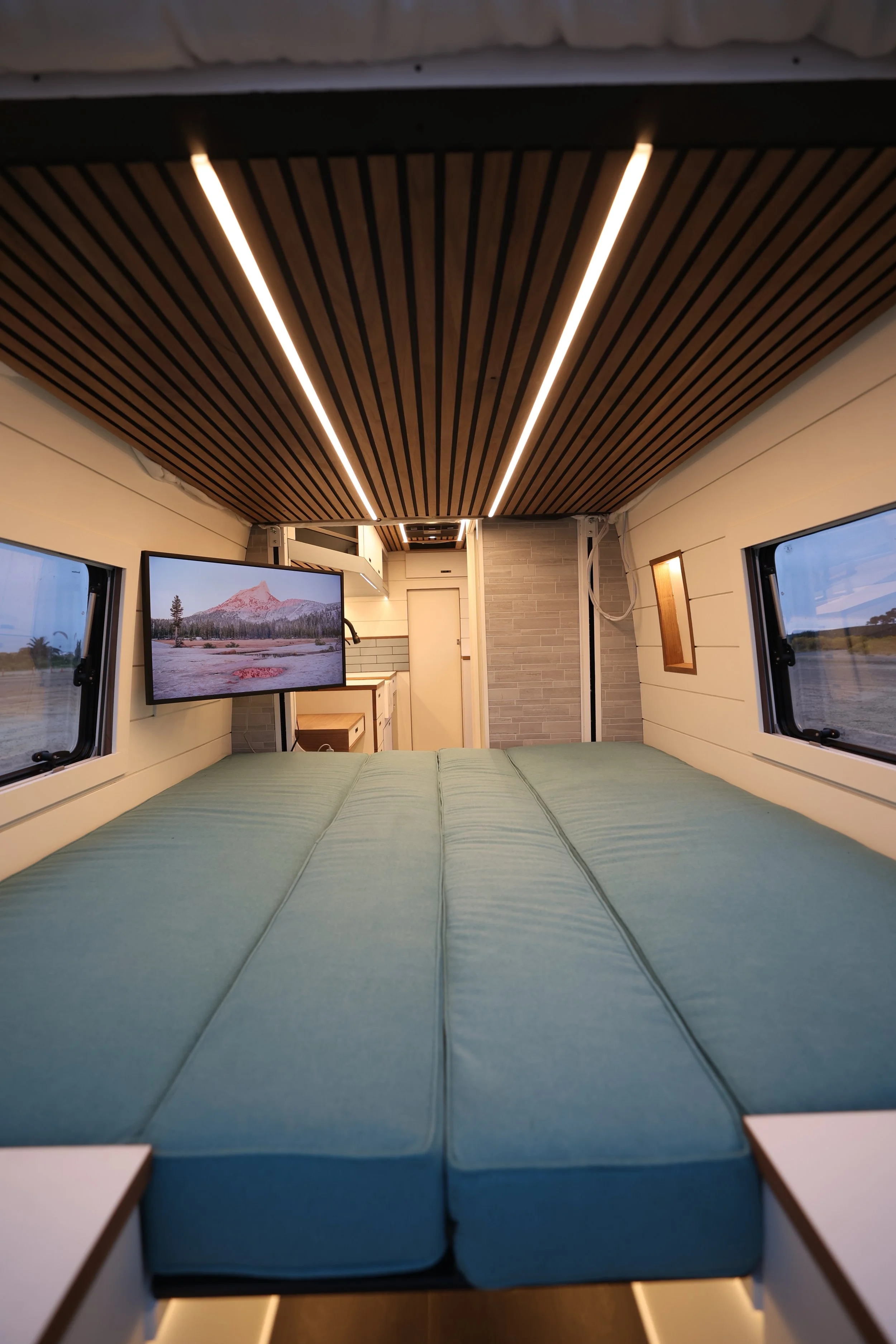 Interior of a compact camper van with a bed, two windows, a television, and modern decor.