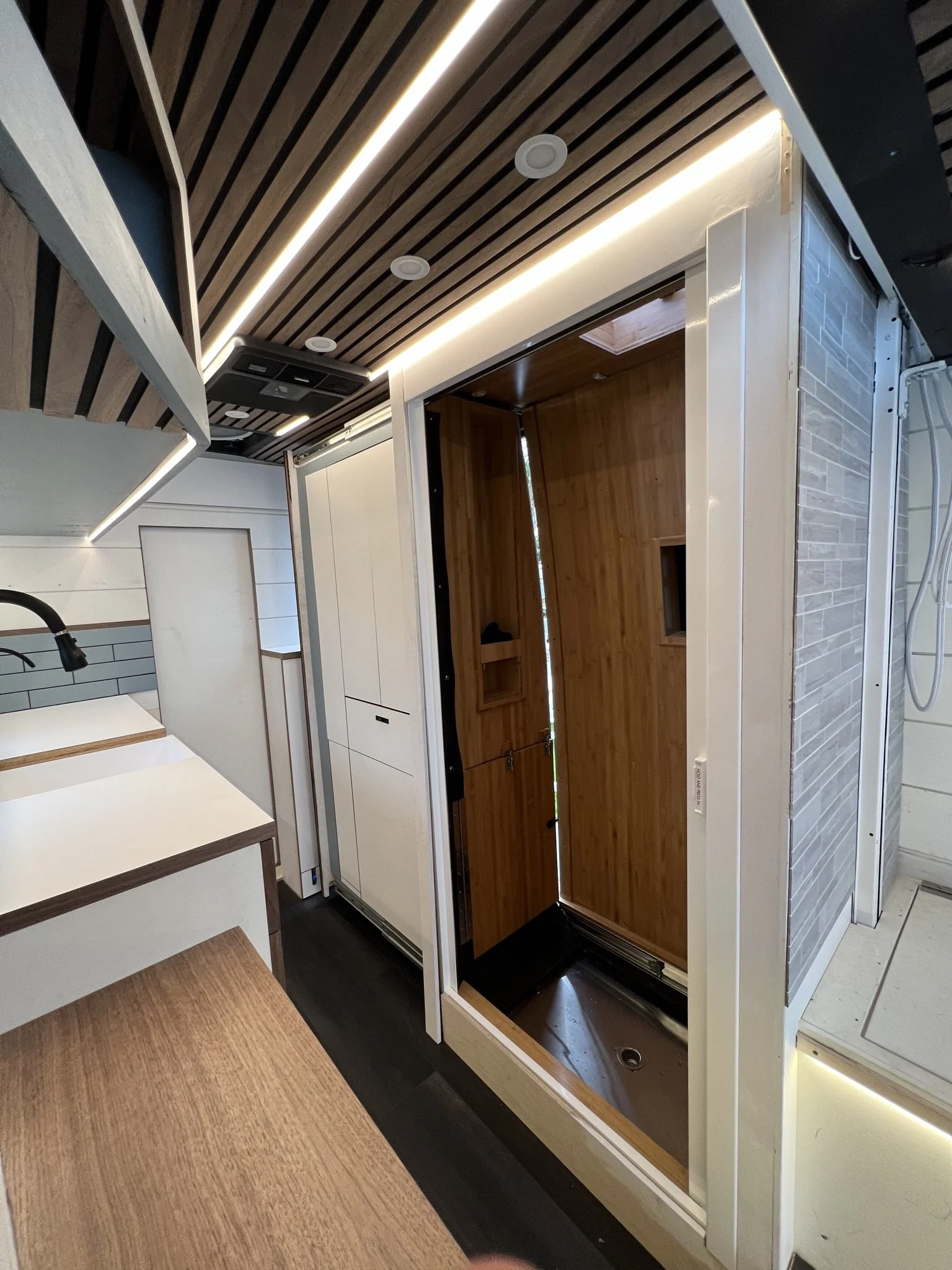 Interior of a small bathroom or shower room with a wooden shower enclosure, white framing, and modern lighting. The room has a wooden ceiling and a nearby sink with a black faucet.