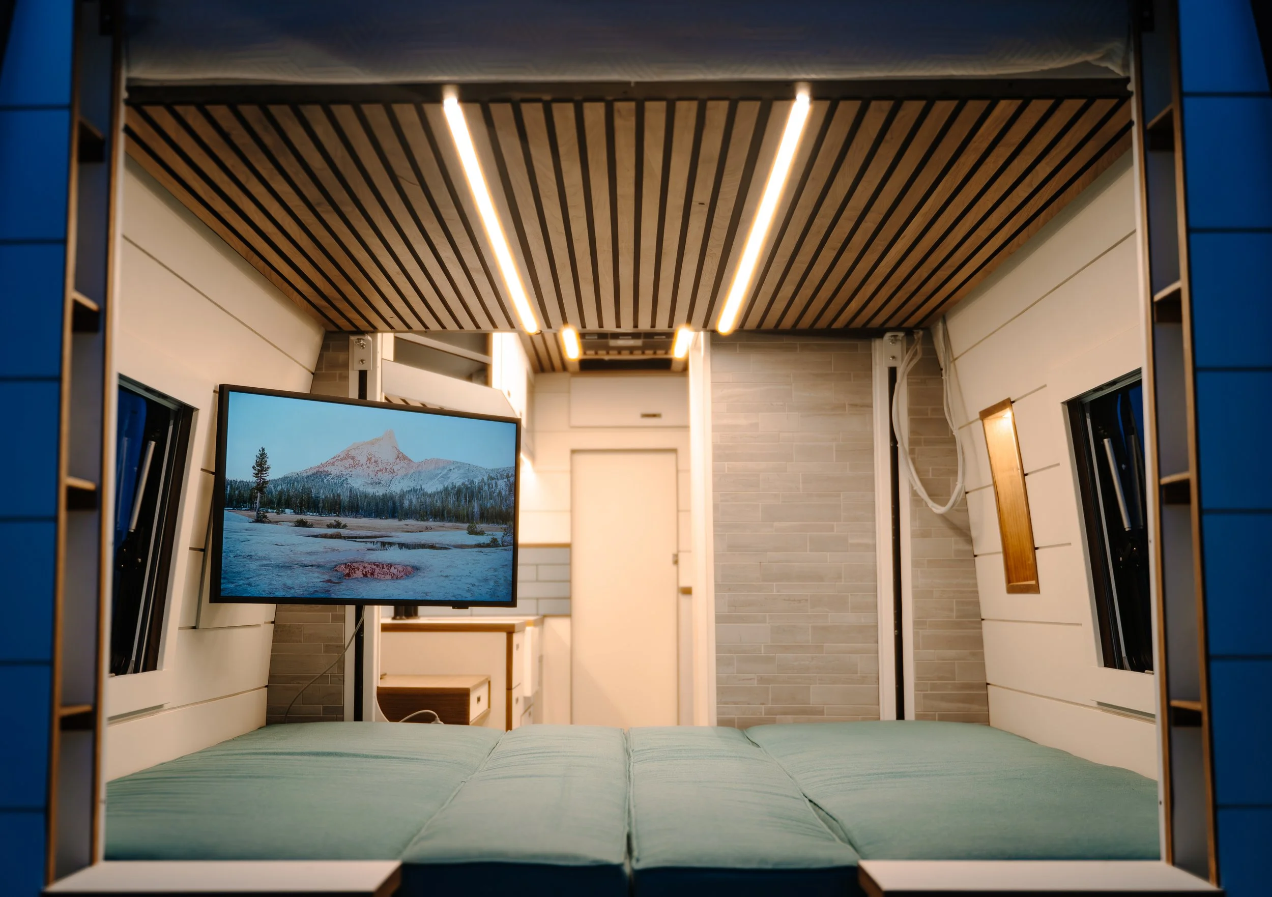 Interior of a tiny home or camper with a bed, a mounted television showing a mountain scene, and small windows on the sides.