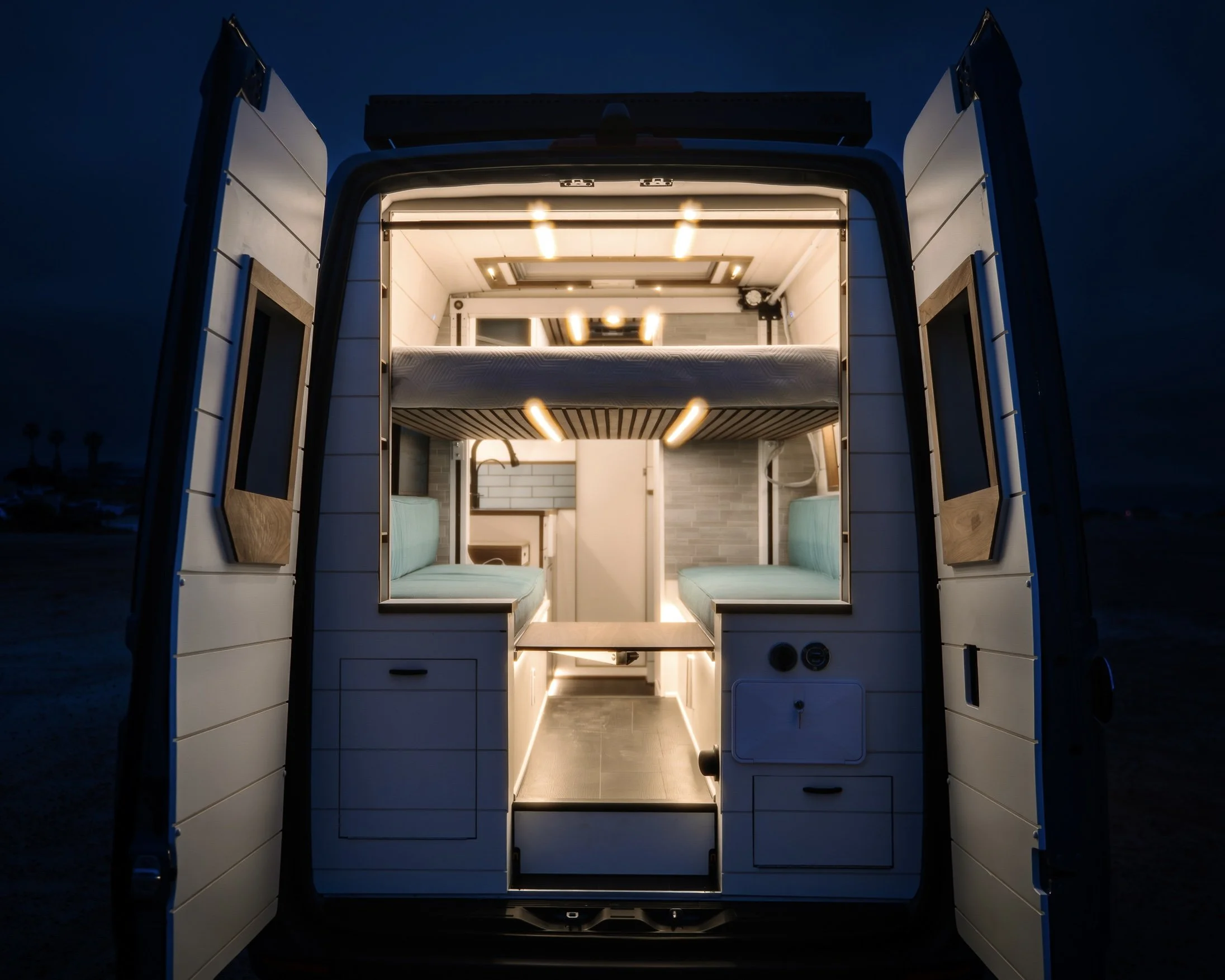 Interior of a converted camper van with built-in seating, a bed, and kitchen appliances, illuminated at night.