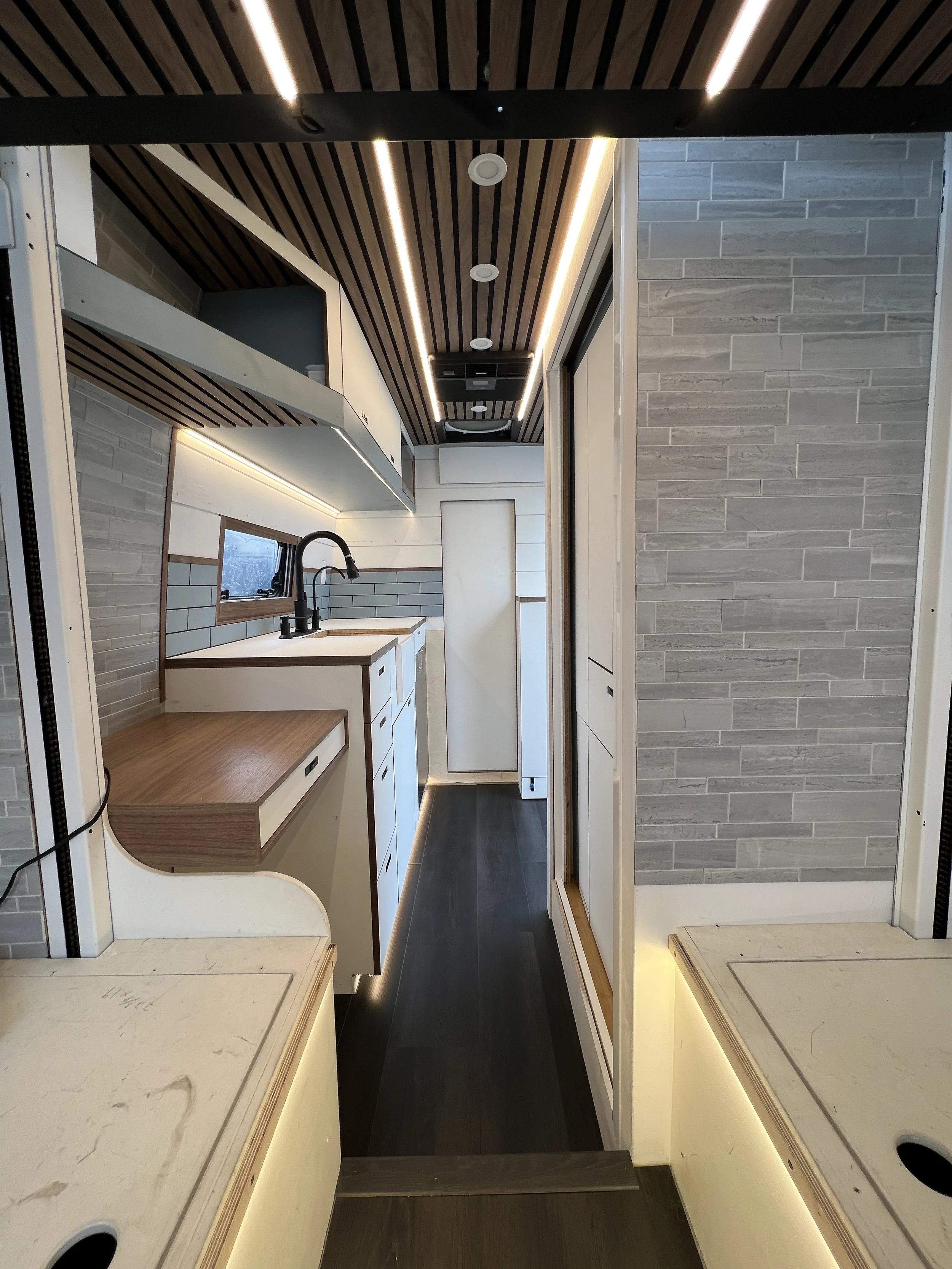 Interior view of a modern tiny home or camper kitchen with white cabinets, a black faucet, a small window, wood accents, and stylish lighting.