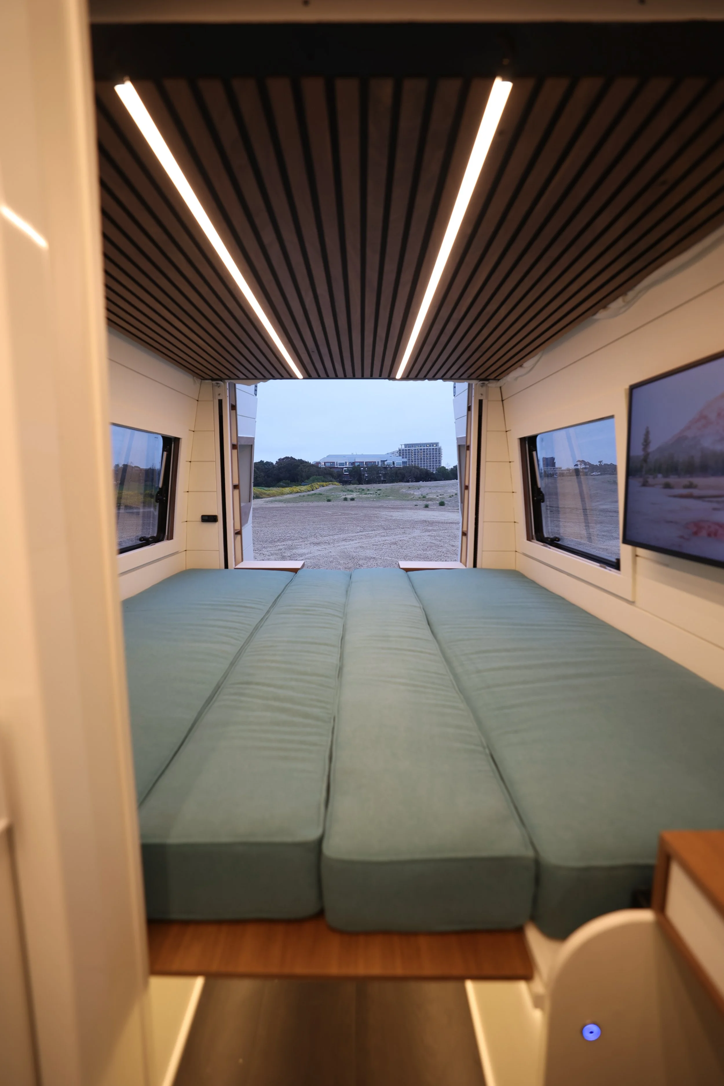 Interior of a small, cozy camper with a bed made of three mattresses, windows on each side, and an open back view showing an outdoor landscape with a few buildings and trees.