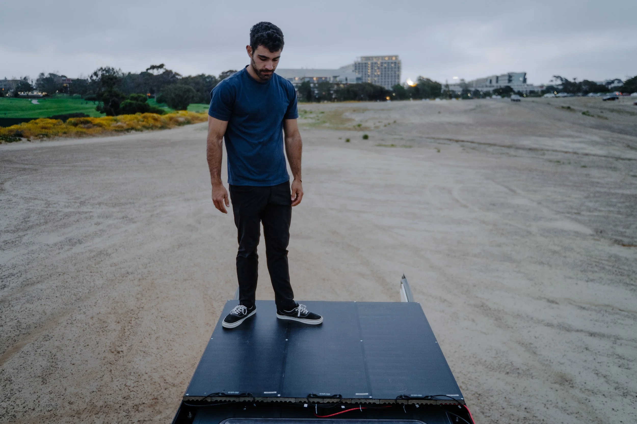 Young man standing on a solar panel on a sandy beach, looking down with an overcast sky in the background.