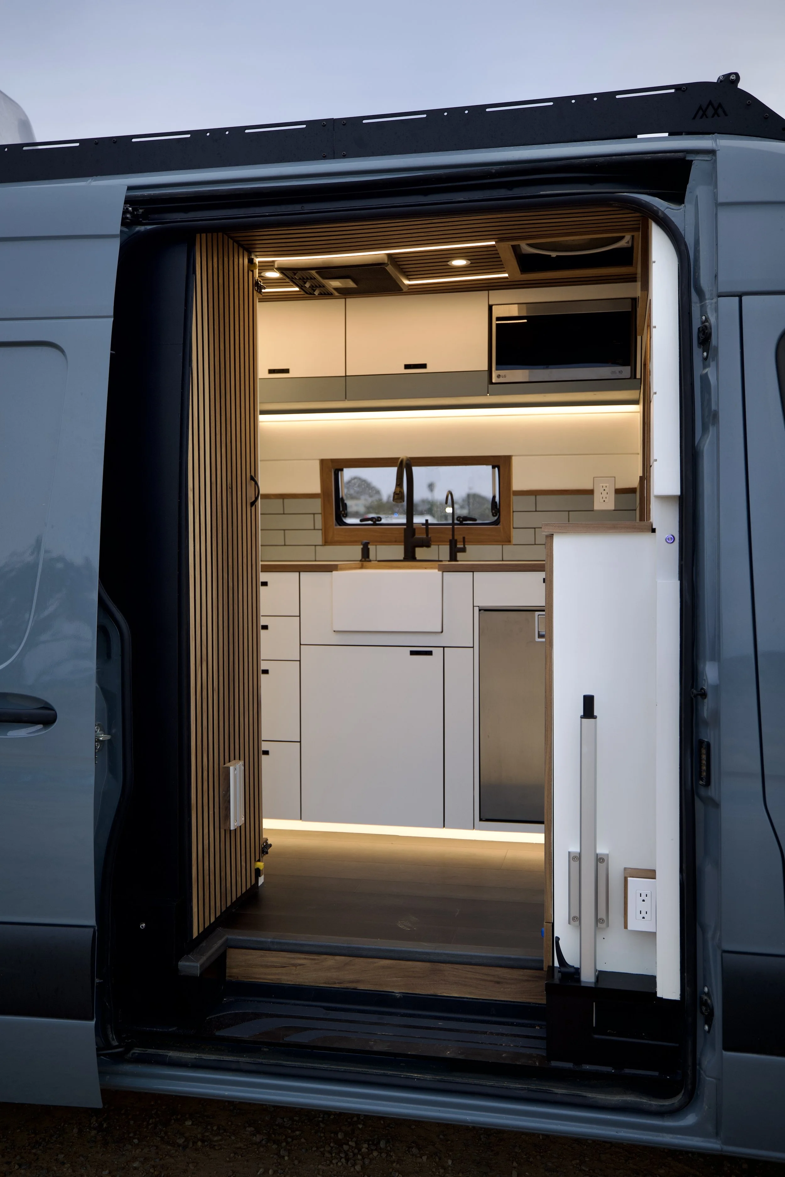 Interior of a modern, compact camper van kitchen with white cabinets, a black faucet, a microwave, and a small window, illuminated with warm LED lighting.