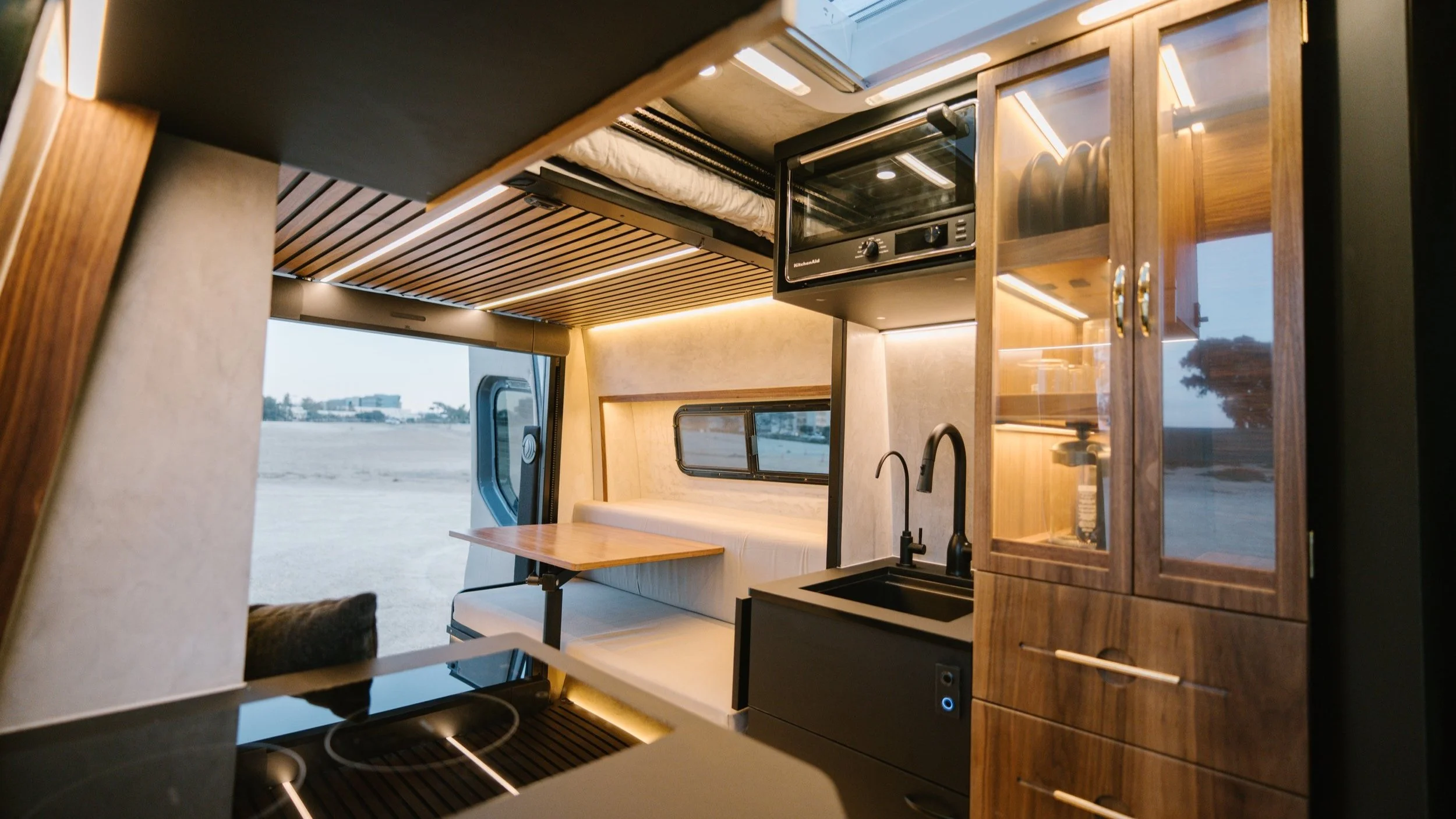 Interior of a compact camper van with wooden cabinetry, a small picnic table, a window with a view of the outdoors, a black sink, and an oven mounted on top of the wooden cabinets.