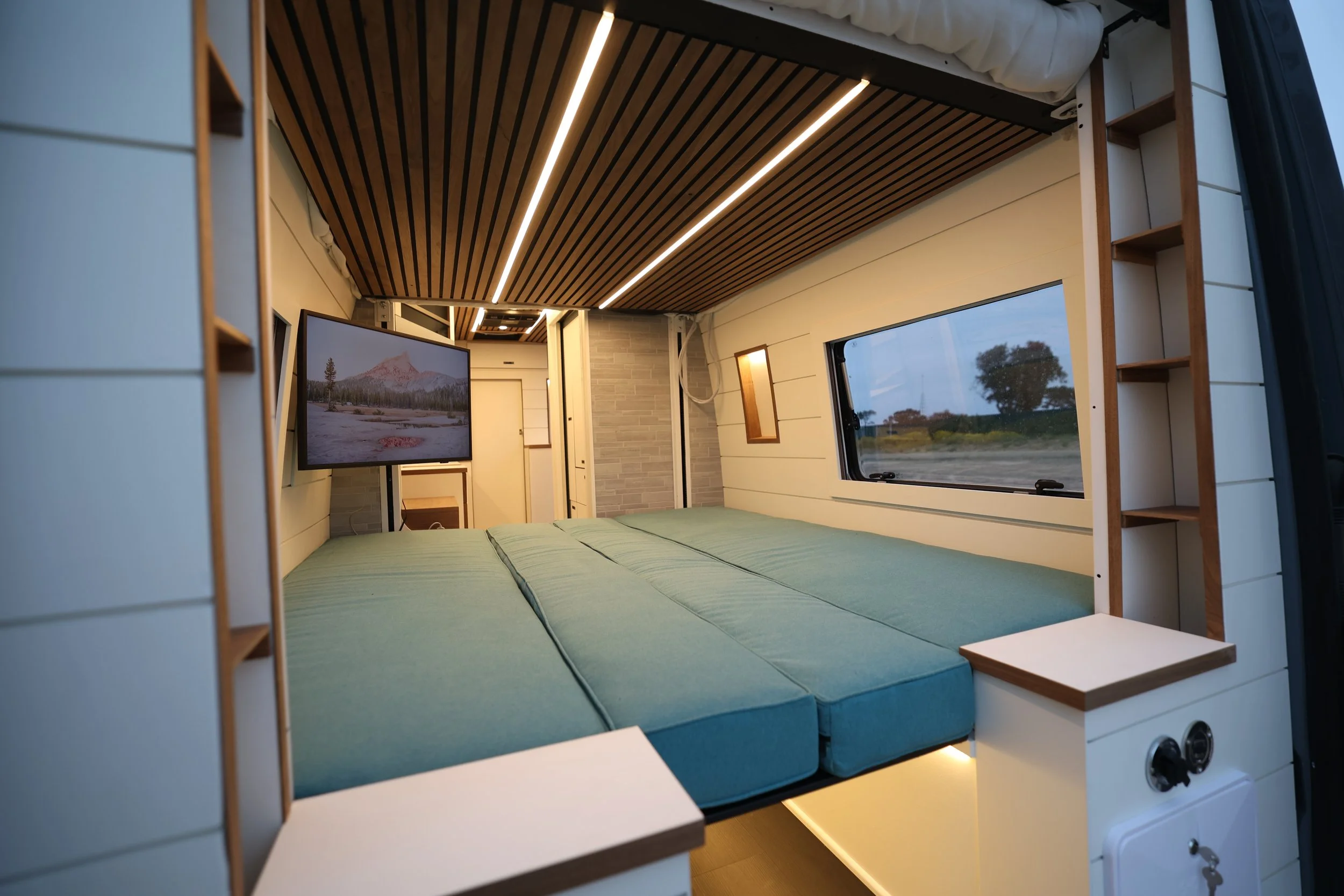Interior of a compact modern camper van with a bed, large window, and overhead TV, featuring wooden and white panel accents and LED lighting.